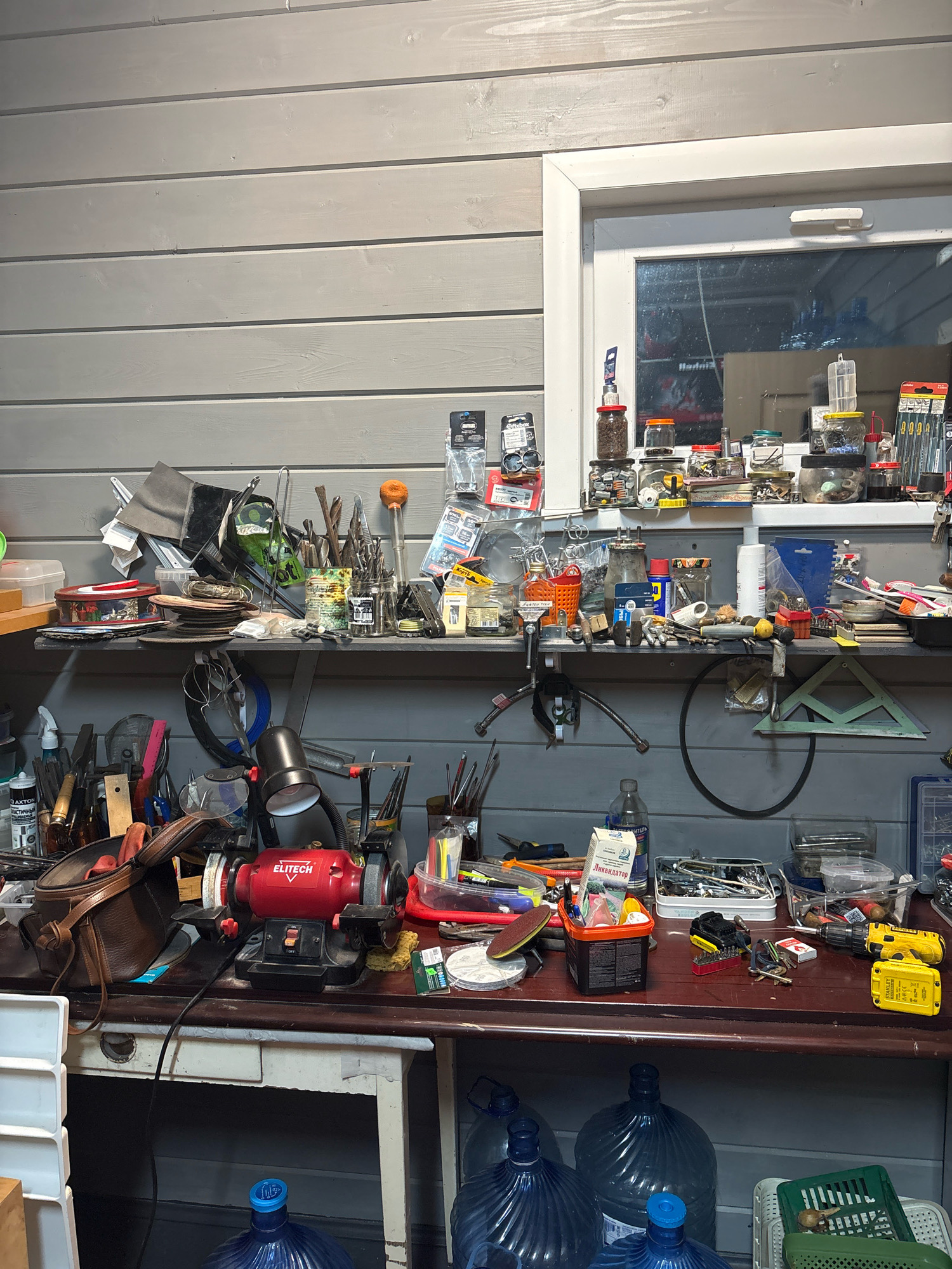 The image depicts a cluttered workshop with a wooden planked wall and a small window. A workbench is visible, covered with various tools and supplies. On the workbench, there is a red electric grinder with the brand "ELITECH" and a brown leather bag. A shelf above the workbench is also filled with jars containing small items, tools, and other miscellaneous objects. In the foreground, several large plastic water bottles are visible, along with some storage containers. The lighting suggests it is daytime, possibly indoors with artificial lighting. There are no people or animals in the image. The overall impression is of a functional, albeit disorganized, workspace.