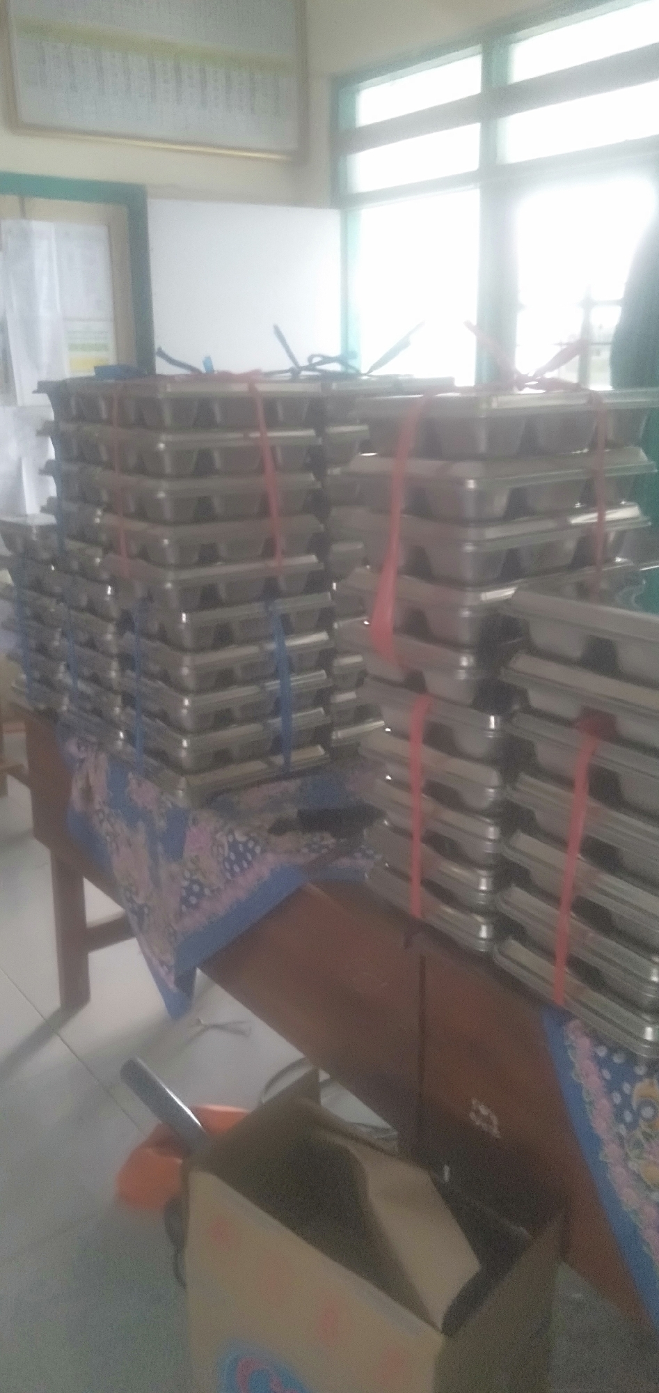 The image displays stacks of identical metal trays, possibly for food service or storage, arranged on a wooden table. The trays are secured with red and blue plastic ties. A patterned fabric, blue with pink and white floral designs, is draped over the table. In the background, large windows are visible, letting in bright light that washes out some details. A framed chart or poster hangs on the wall above and to the left of the trays. To the right, a person's shoulder and arm are partially visible, wearing dark clothing. On the floor in the lower foreground, a partially opened cardboard box and a broom are present. The scene appears to be indoors, possibly in a school or community hall setting. This image is not geographically specific to Plemahan, Indonesia, and no landmarks or context suggest that location.