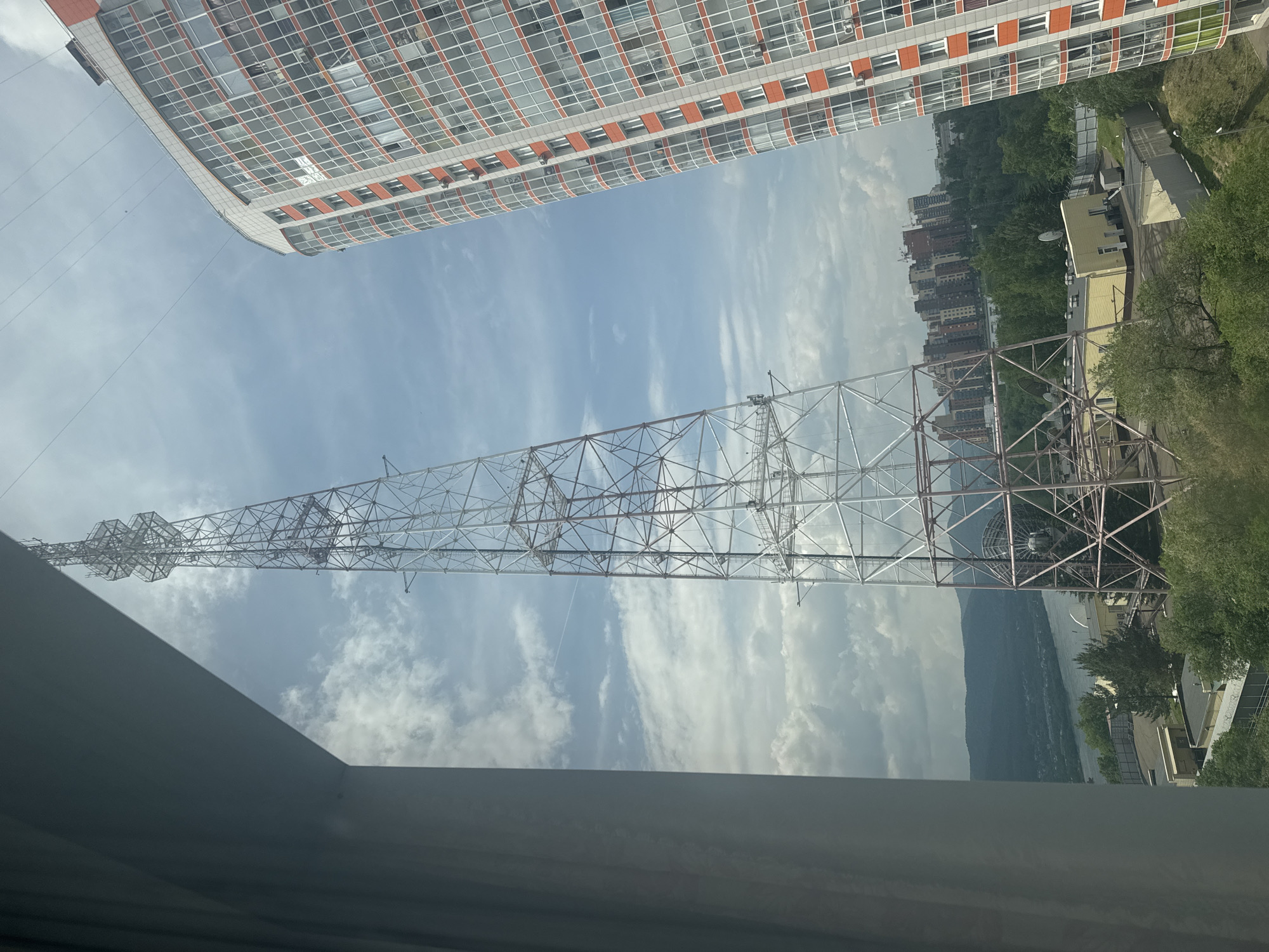 The image shows a view from a window, looking out at a telecommunications tower, a modern residential building, and a distant cityscape with hills and a river. The telecommunications tower, a tall lattice structure, dominates the center of the frame. To its right is a multi-story residential building with a curved facade, featuring numerous windows and orange accents. In the background, a collection of taller buildings can be seen, indicative of a city center. A wide river or body of water is visible in the distance, bordered by lush green hills. The sky is partly cloudy, with some distinct cloud formations. The scene is likely located in the city of Krasnoyarsk, Russia, given the distinctive geography featuring a large river (likely the Yenisei) and surrounding hills. There are no people visible in the image. The overall impression is an urban landscape with industrial and residential elements set against a natural backdrop.