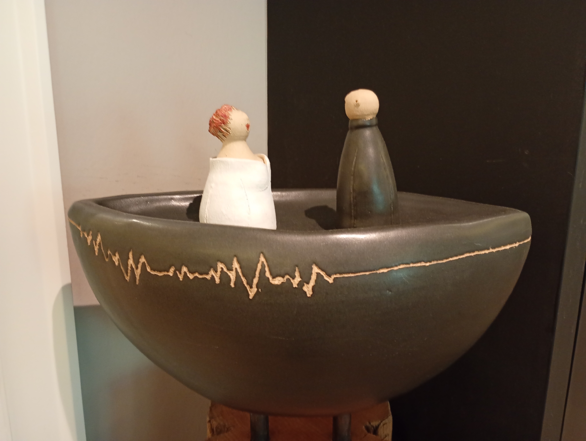 The image shows a close-up of a dark, wide, ceramic bowl with a textured, light-colored wavy line design around its rim. Inside the bowl are two small, stylized figurines. The figurine on the left has a light-colored body with red hair and is wearing a white garment that covers its lower body. The figurine on the right is dark, conical, and has a light-colored head. The bowl is resting on a rough, wooden base. The background is divided vertically into a light gray-colored wall on the left and a dark gray to black wall on the right. The scene appears to be an indoor display, possibly in a museum or gallery.