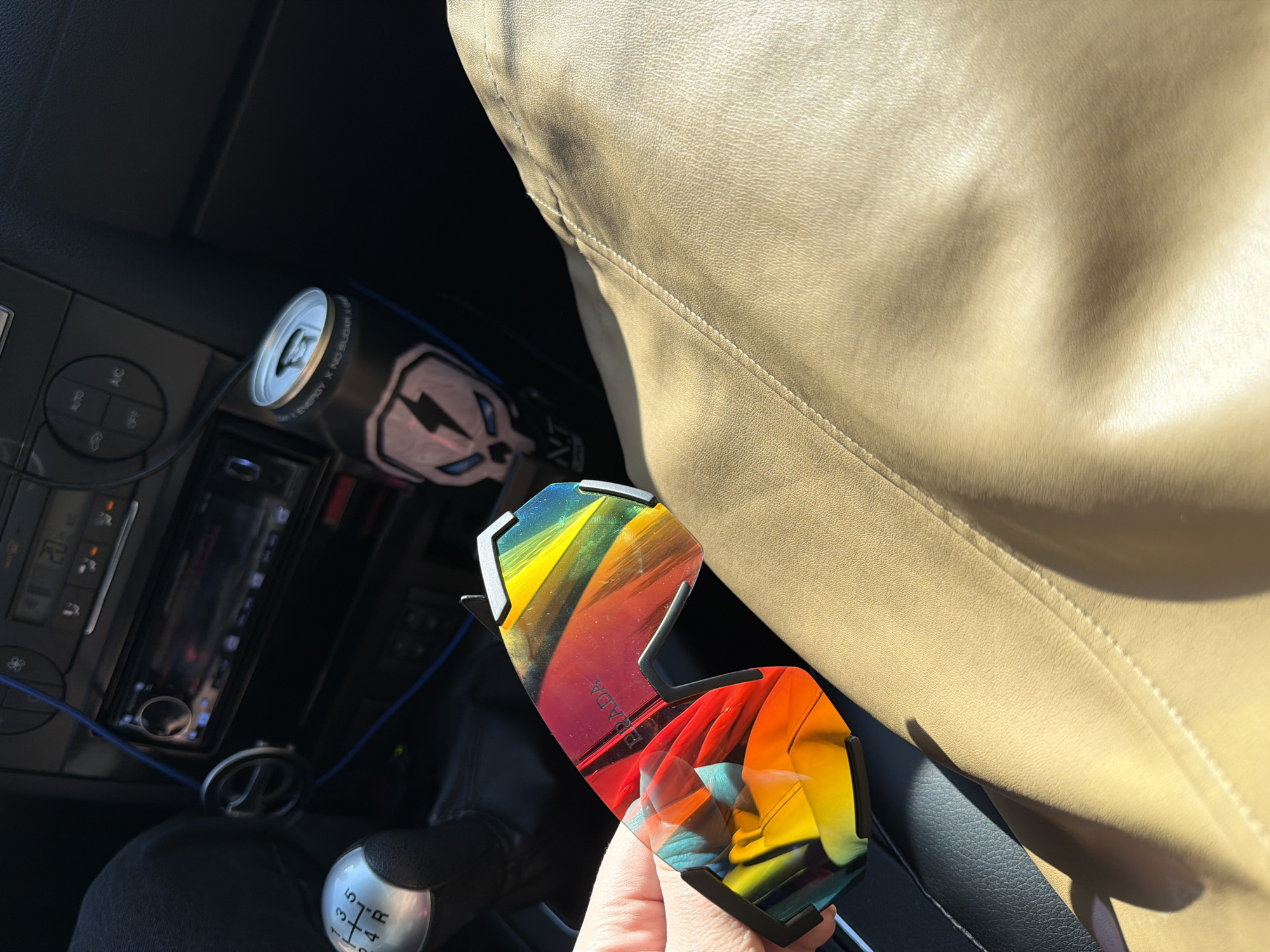 The image shows the interior of a car. A hand is holding a pair of colorful Prada sunglasses. Next to the sunglasses is a tan leather seat. In the background, on the center console, there is a can of energy drink and the car's radio/CD player. The gear shift is visible in the lower left. I have no information that indicates that it is in Chaikovsky, Russia.