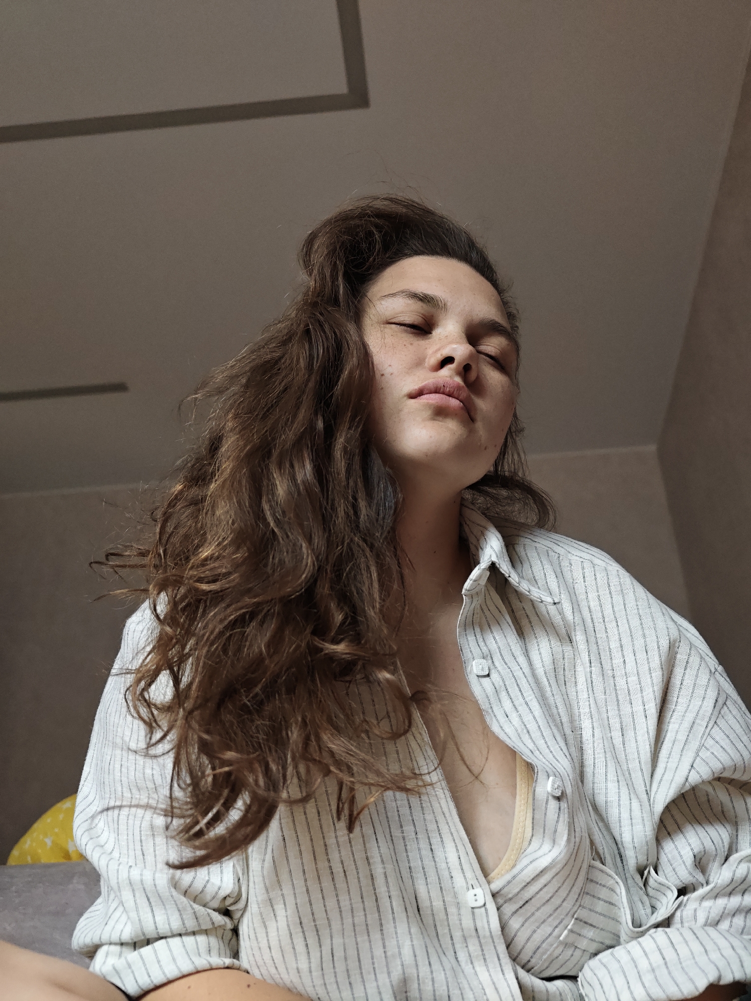 A low-angle, medium shot depicts a fair-skinned young woman with her eyes closed. Freckles are visible on her nose and cheeks. Her wavy, brunette hair is styled with some pulled back on top of her head. She is wearing a vertically striped white and grey button-down shirt, unbuttoned enough to reveal a glimpse of a light yellow bra.
The background is out of focus. The upper portion of the shot shows an off-white ceiling with a darker, rectangular recessed area. A soft beige wall is visible in the background.
The location is presumed to be indoors, possibly in a home, in the city of Chaikovsky, Russia. No other details about the location or the woman's actions are ascertainable from the image.