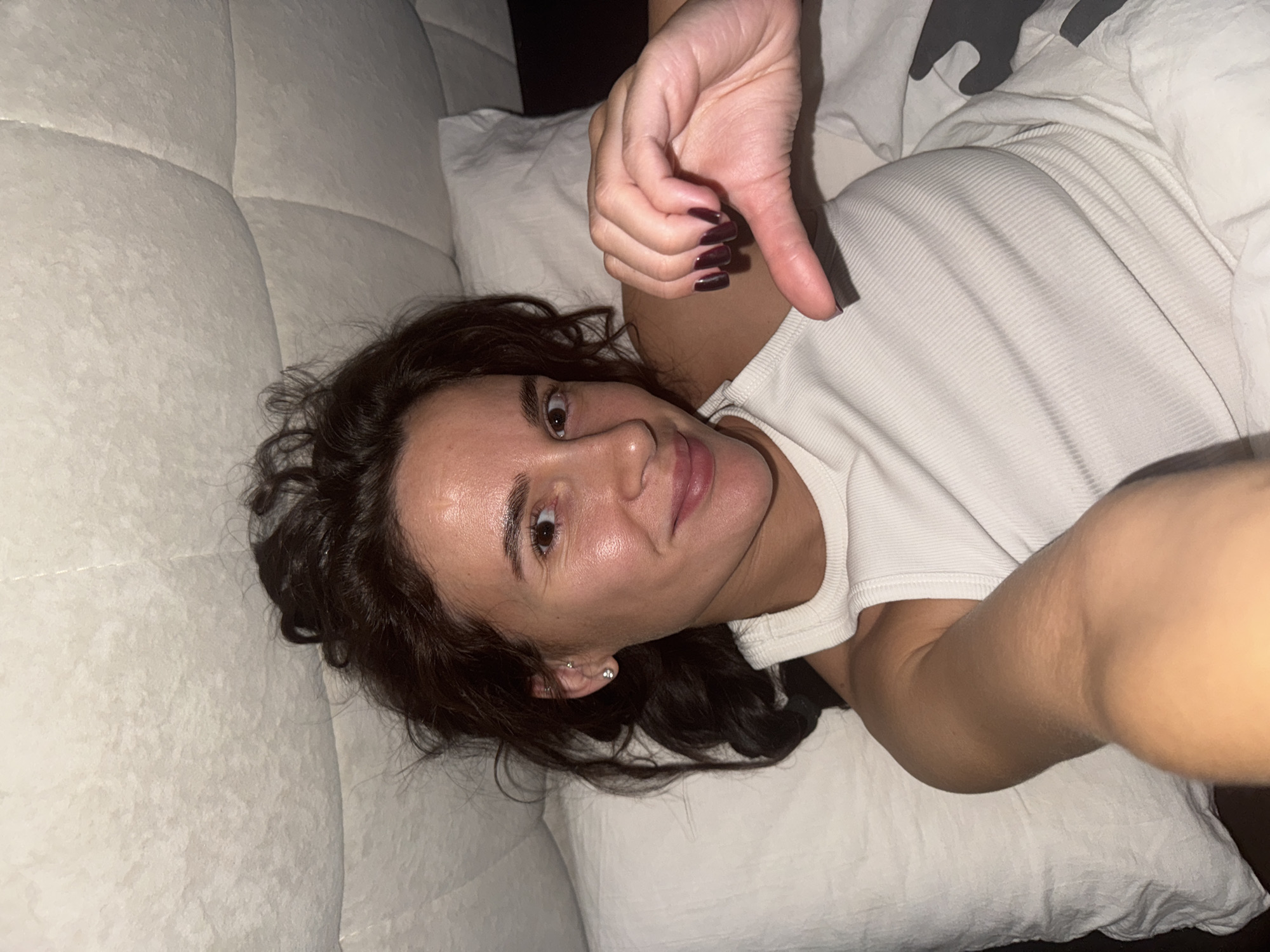 A woman is lying on a bed covered in white linens and a white, ribbed tank top. She is giving a thumbs-down gesture with her left hand, which has dark red nail polish. Her dark brown hair is loose and slightly disheveled. The background is blurred. There is no indication of Chaikovsky, Russia.