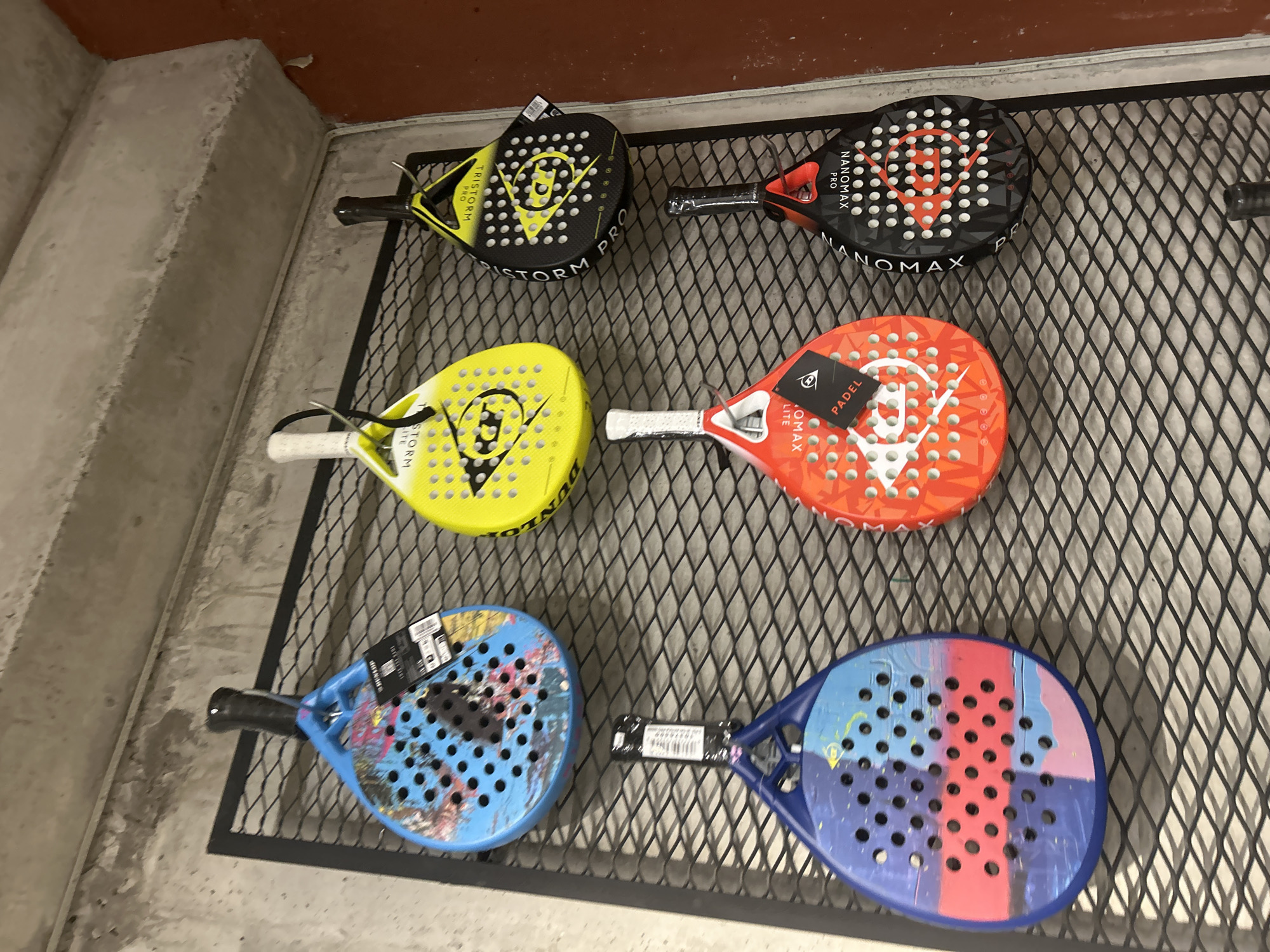 Six padel rackets are displayed on a black metal grid against a concrete wall. The rackets are arranged in two rows of three. From left to right, top row: a blue and pink patterned racket with a world map design, a yellow racket with a black Dunlop logo, and a black and yellow racket with "TRISTORM PRO" written on it. Bottom row: a blue and pink patterned racket, a red racket with "PADEL" and "NANOMAX LITE" written on it, and a black racket with "NANOMAX PRO" written on it. All rackets are hanging from hooks attached to the grid. The background shows a textured concrete wall and a narrow band of reddish-brown wall. The lighting is bright, casting shadows from the rackets onto the grid. The image appears to be taken in an indoor retail setting, possibly a sports equipment store. The location is identified as being in Tbilisi, Georgia.