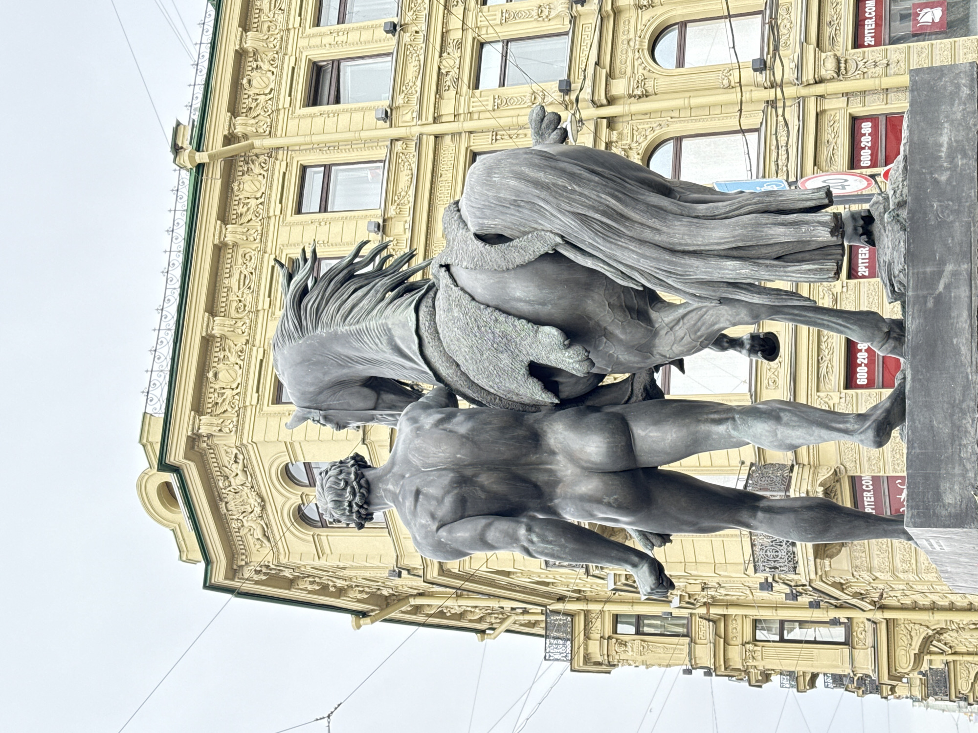 A bronze statue of a nude male figure wrestling a rearing horse stands in front of a yellow building in Saint Petersburg, Russia. The statue is positioned on a dark gray plinth. The building behind the statue has a highly decorated facade with multiple windows. Signs with the text "2PITER.COM" and the number "600-20-80" are visible on the building. Wires are strung across the scene. The sky is overcast.