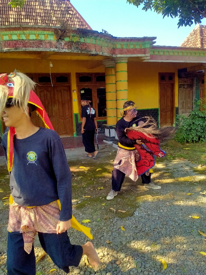 The image depicts an outdoor scene in Tanon, Indonesia, featuring three males in what appears to be performance or ceremonial attire in front of a residential building under clear daylight.

In the immediate foreground, to the left, a male subject is partially visible, facing left and smiling slightly. He wears a dark long-sleeved shirt with a circular emblem, a patterned light-colored sarong-like garment, dark trousers, and is barefoot. His head is adorned with a large, elaborate red and yellow headpiece with blonde/white hair-like strands and dark sunglasses.

In the mid-ground, to the right, a second male subject stands with his legs spread, facing right. He wears a dark long-sleeved shirt, a yellow headband, and has dark face paint lines on his cheeks. He also wears a patterned light-colored sarong-like garment over dark trousers and light-colored shoes. He is holding a large, red, intricately decorated object with white and black accents, from which brown, straw-like material protrudes.

Further back, in the center, a third male subject walks towards the right. He wears a dark short-sleeved t-shirt with white text, dark trousers, a dark baseball cap, and light-colored footwear. He holds a thin, dark, stick-like object in his right hand.

The background features a single-story building with bright yellow walls, multiple brown wooden doors, and decorative green and yellow columns. The roof is constructed of brown tiles, and the roofline includes a concrete trim that shows signs of weathering and minor plant growth. The ground in the foreground and mid-ground is a mix of loose gravel, dirt, and patches of green moss or grass, with scattered dry yellow leaves. The overall lighting suggests a sunny day, casting discernible shadows on the ground.
