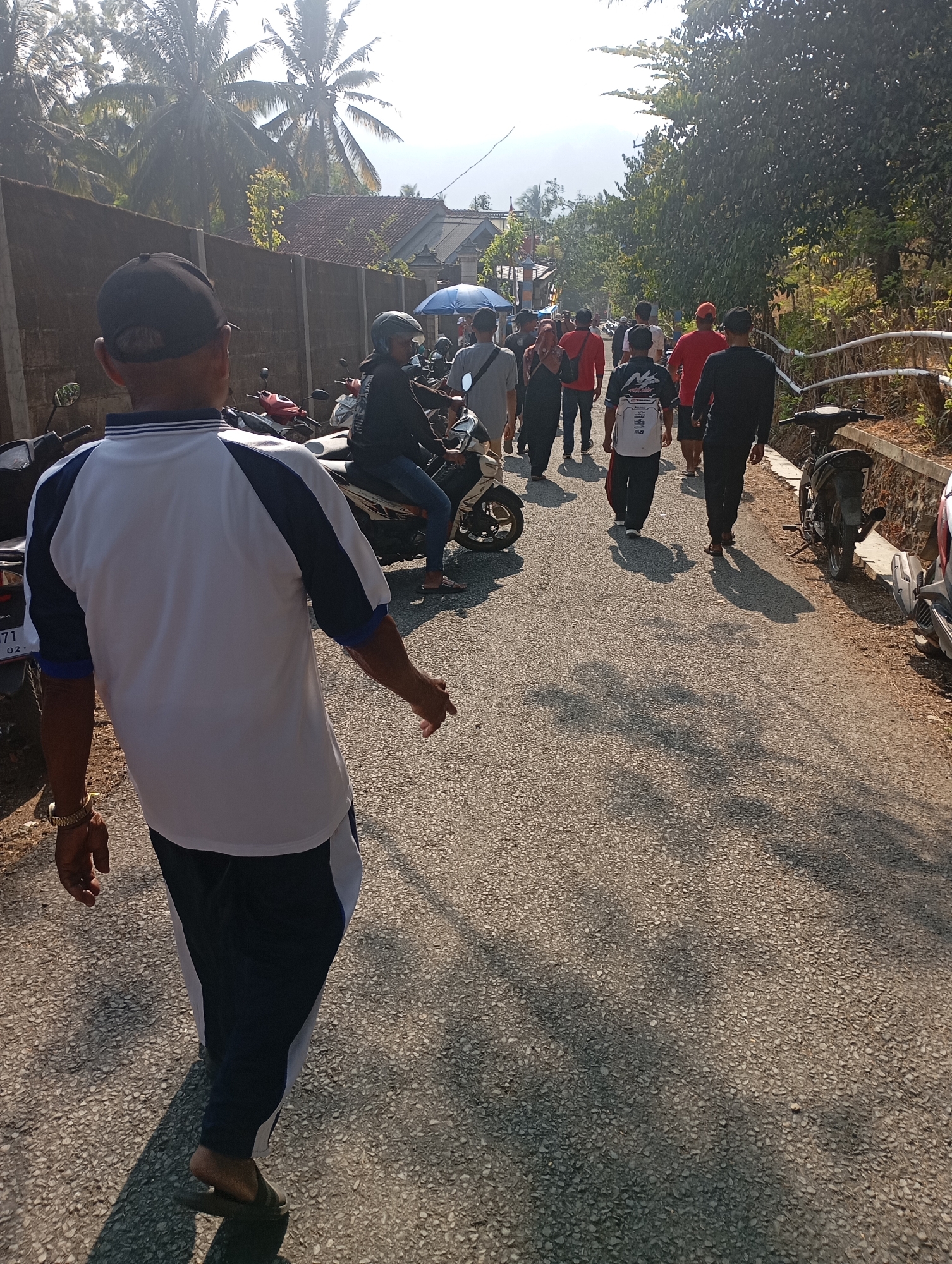 The image depicts a street scene in Ketepung, Indonesia, on a sunny day. In the foreground, a person is seen from behind, wearing a white and blue short-sleeved shirt and dark pants. They are walking away from the camera, casting a shadow on the asphalt road. The road is lined with trees on the right, and a wall on the left. Several other people are walking in the same direction on the road, some appearing to be part of a group. A person is riding a motorcycle on the left side of the road, and a few other motorcycles are parked along the side. The environment suggests a rural or semi-urban setting with residential structures visible in the background.