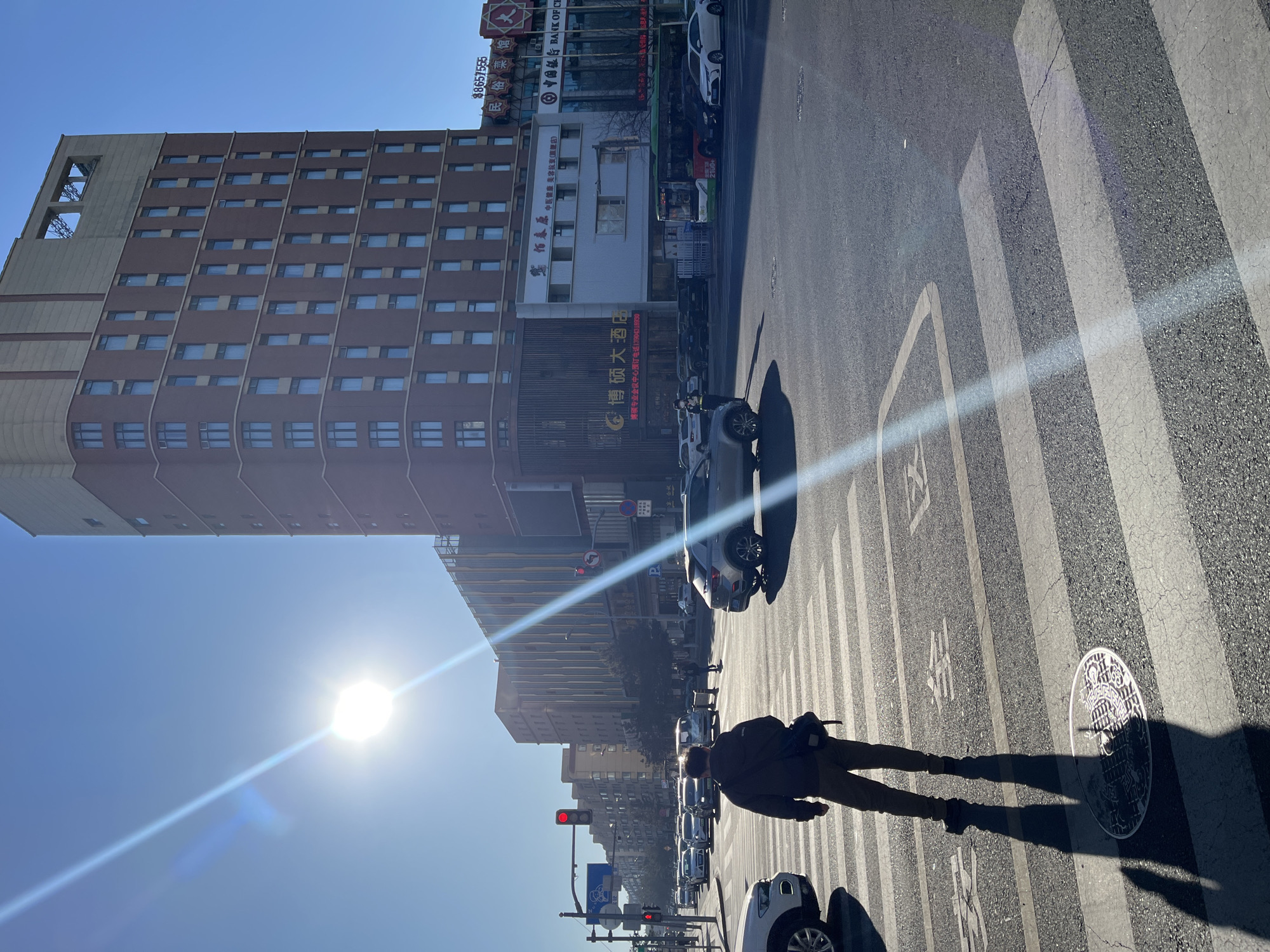 The image shows a street scene in Changchun City, China on a sunny day. A person walks across a zebra-striped crosswalk with their shadow stretched out on the pavement. The street is lined with cars and buildings. The sun shines brightly in the sky, creating a lens flare. Signs for businesses such as the "Bank of China" and "Bo Suo Grand Hotel" can be seen on the buildings. Traffic lights are visible in the distance. The environment appears to be an urban area with typical street infrastructure and a mix of commercial and residential buildings.