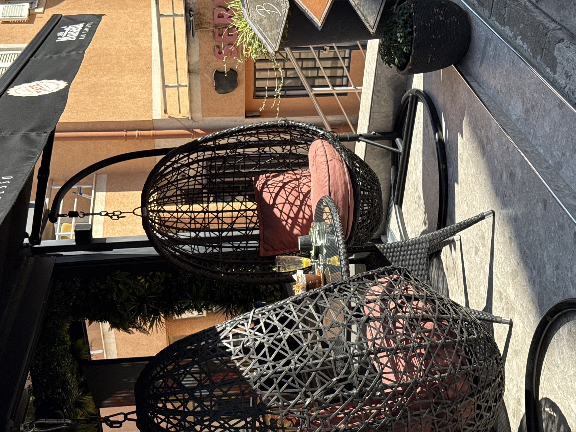 An outdoor seating area of a cafe in Budva, Montenegro, features two hanging egg chairs with reddish-pink cushions and a small glass-top table. On the table, a partially consumed drink in a tall glass sits next to a small brown paper cup filled with snacks and an ashtray. A potted plant is visible to the right of the chairs. The cafe's black awning, bearing a logo that reads "TATE", extends over the seating area. In the background, an orange building with white-shuttered windows and a metal railing is visible, suggesting a street-level or elevated cafe setting. The scene is illuminated by bright sunlight, casting shadows on the tiled floor.