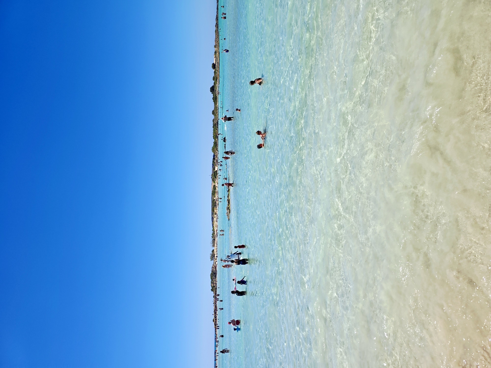 The image depicts a view of a crowded beach with clear, turquoise water and a bright blue sky. Numerous people are visible in the shallow water, some swimming, others standing or wading. The water is transparent, revealing the sandy bottom. In the distance, a thin strip of land with vegetation and some structures can be seen, suggesting a coastline. The overall scene is characteristic of a popular beach destination during daylight hours, likely in a Mediterranean climate. The location is identified as Elafonissi Beach, Crete, Greece.