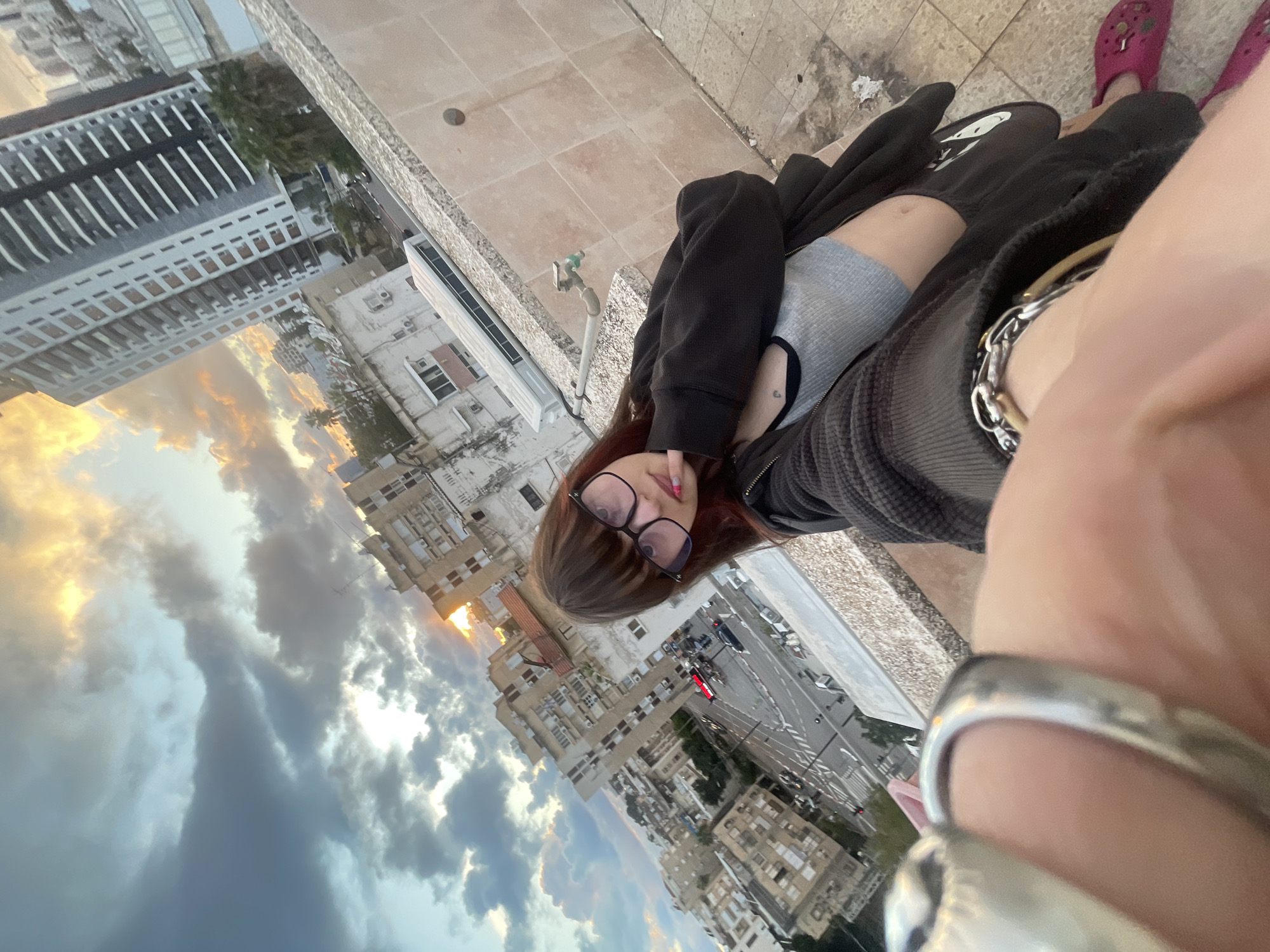 The image depicts a high-angle selfie taken from a rooftop or elevated balcony overlooking a city street in Petah Tikva, Israel, during what appears to be sunset or twilight due to the dramatic sky.
**Subject:** A young person, appearing female, with dark brown hair, bangs, and wearing dark-framed eyeglasses, is positioned centrally. They are dressed in a light grey cropped tank top, an open dark hooded jacket, and dark bottoms. Bright pink clogs are visible on their feet. A small dark tattoo, possibly a heart, is visible on their chest. Their right hand, adorned with a large silver-toned ring on the ring finger and a silver chain bracelet on the wrist, holds the camera to capture the selfie. Their left hand is raised, with the index finger touching their chin. Nails on the left hand display colorful polish.
**Environment and Background:** The immediate foreground features a tiled rooftop surface (light brown/beige square tiles) and a low, light-colored granite or marble railing/wall. A metal spigot with a green handle is affixed to this wall. Beyond the railing, a bustling urban landscape unfolds. A multi-lane street with vehicles, including a red car at a traffic light, is visible below, along with white crosswalk markings. Various multi-story buildings populate the background: an older, light-colored apartment block with air conditioning units to the left, and a taller, more modern high-rise building with a grey and white facade to the right. The sky overhead is partially cloudy, with dark grey clouds interspersed with bright patches of golden-orange light from the setting sun. Distant trees and other urban structures are visible throughout the city vista.