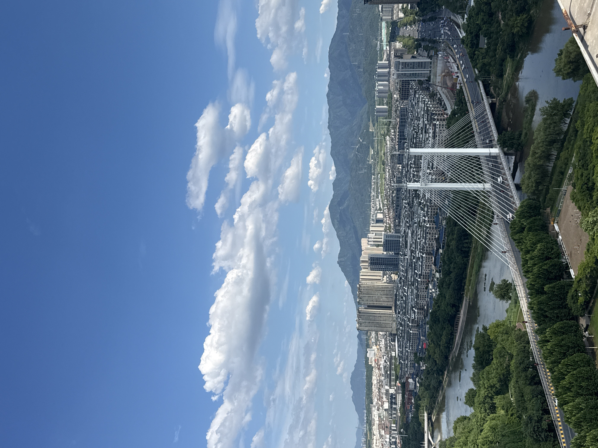 This is a high-angle drone shot capturing an aerial view of a city in Yiwu, China, on a clear day with scattered white clouds. The scene is dominated by a wide river flowing through the urban landscape. A prominent cable-stayed bridge with two white towers spans the river, with vehicles moving across it. Beyond the river and bridge, a densely packed residential area with numerous buildings, many appearing to be multi-story apartment complexes, stretches towards a backdrop of lush, green mountains. The mountains are partially obscured by a haze. The sky is a vibrant blue, with cumulus clouds casting shadows on the terrain below. The overall impression is one of a modern, developing city integrated with its natural surroundings.
