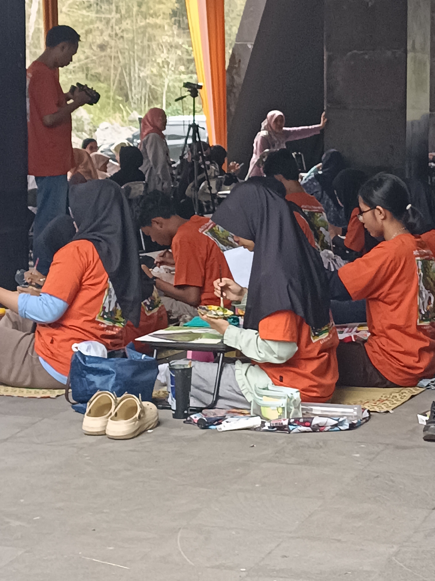 In Hargobinangun, Indonesia, a group of people are gathered outdoors, possibly for an art workshop or event. In the foreground, two pairs of casual sandals are placed on the concrete ground. Several individuals, mostly young women wearing hijabs, are seated on the ground, engaged in painting. They are working on canvases laid out on portable tables, with various art supplies like brushes and paint palettes visible. One person, wearing an orange t-shirt and blue jeans, stands on the left side of the frame, holding a camera and appearing to film or photograph the activity. In the background, more people are seated, and some are standing. The setting features some greenery and bamboo-like plants, with orange fabric draped in the background, suggesting an organized event possibly under a temporary shelter or open structure.