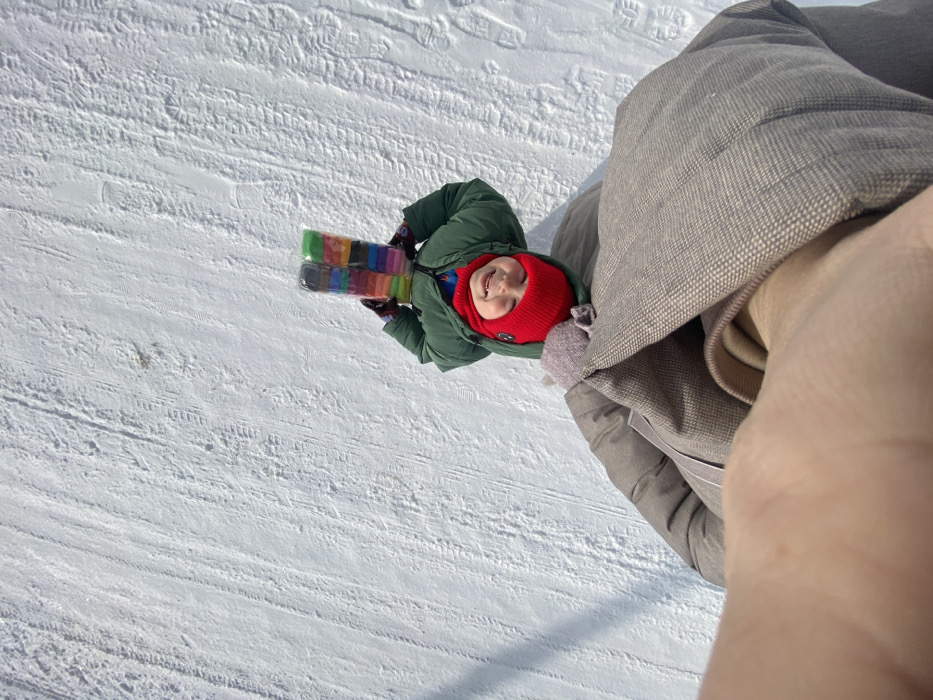 This image, taken in Узяр, Russia, captures a scene from a high-angle perspective, looking down onto a snow-covered outdoor environment. In the center, a young individual, likely a child, is visible, wearing a dark green winter jacket, a bright red knit beanie, and red or burgundy gloves. The child's face is smiling and directed upwards towards the camera. Both hands are raised, holding two transparent plastic packages containing multiple rectangular blocks of colorful modeling clay or craft dough. One package has a prominent green top, and the other a darker grey or black top. The ground beneath the child is entirely covered in white snow, displaying various tire tracks and scattered footprints. The bottom portion of the frame shows parts of a person's light brown or beige winter jacket and a hand, indicating the camera is being held by that individual. The lighting suggests a clear, sunny day.