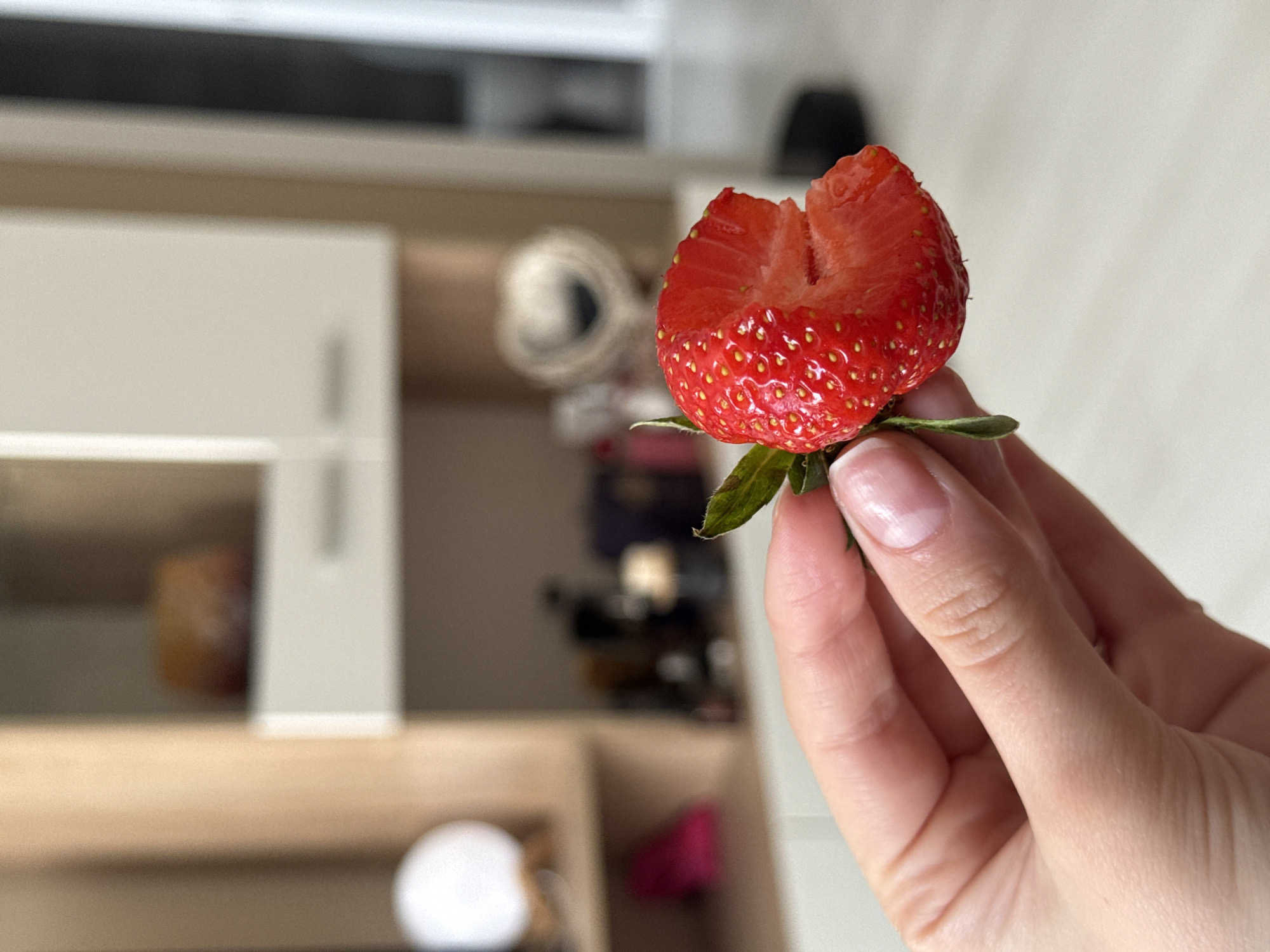 A close-up shot features a human hand holding a bright red strawberry, which appears to be cut in half or partially eaten. The strawberry is the main subject, sharply in focus, displaying its vibrant red flesh, tiny yellow seeds (achenes) on the surface, and a small green stem attached at the top. The cut side reveals the juicy texture and a lighter, whitish core.
The hand holding the strawberry has fair skin and well-manicured fingernails, some with a natural or light pink polish. The strawberry is held gently between the thumb and index finger, with the other fingers curled slightly.
The background is significantly blurred, creating a shallow depth of field that emphasizes the strawberry and hand. Indistinct shapes and colors in the background suggest an indoor setting, possibly a kitchen or living area. Elements that can be vaguely discerned include light-colored cabinets or shelves, a dark horizontal line at the top, and some blurred items on the shelves, such as a white circular object and a pink-hued object in the lower left. The lighting is bright and even, indicating either natural daylight or good artificial illumination. No text is visible in the image.
