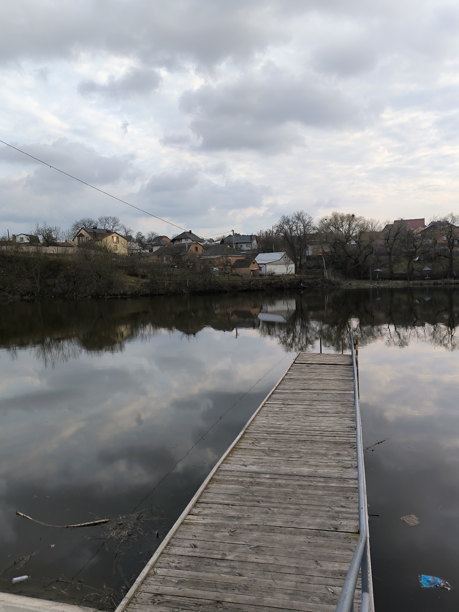 The image depicts a wooden pier extending into a body of water, likely a lake or river, in the city of Vinnytsia, Ukraine. The pier is constructed of weathered planks and features a metal railing on one side. The water surface reflects the cloudy sky above, as well as the trees and buildings lining the opposite shore. The shoreline is visible in the background, with houses interspersed among bare trees. Debris, including plastic refuse and natural materials, is visible in the water near the pier. The sky is overcast with a range of gray and white clouds. A power line stretches across the upper left portion of the image.