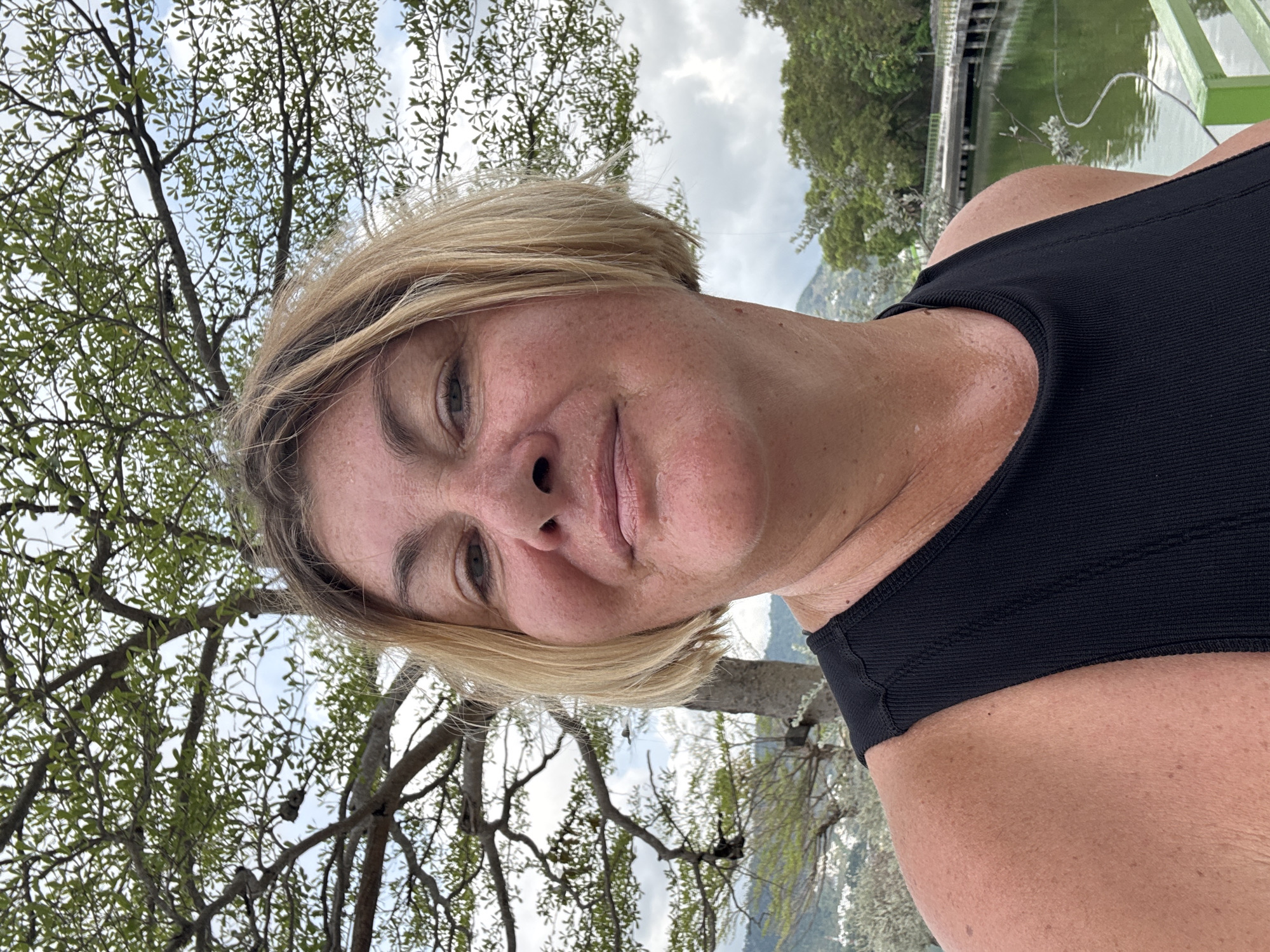 A middle-aged woman with short blonde hair and fair skin is facing the camera in an outdoor setting. She is wearing a black tank top. A tree with sparse green leaves is visible to the left of her face. In the background, there is a body of water, a structure resembling a bridge or dock, and trees, suggesting a park or lakeside environment. Given the prompt's instruction to identify the location as บ้านบางรักษ์, Thailand, it can be inferred that the photograph was taken in a natural setting within that city. There is a hint of mountains in the background.
