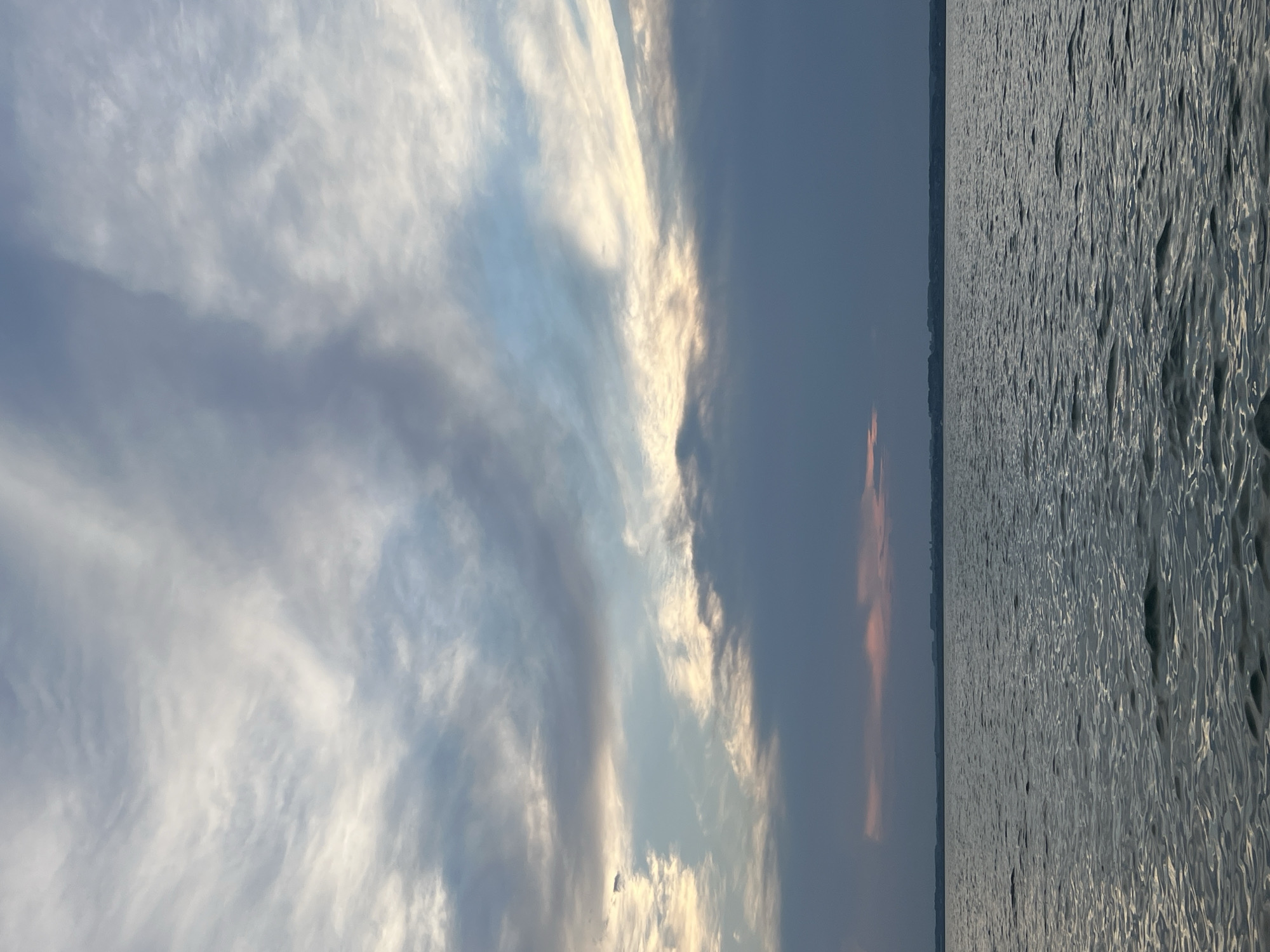 The image depicts a vast body of water under a dramatic sky. The water is a deep gray with gentle ripples reflecting the ambient light. A distant, dark horizon line suggests land with faint structures, possibly buildings or trees. The sky is a canvas of dynamic clouds. Towards the top, wispy white and gray clouds are streaked across a pale blue expanse. Lower in the frame, heavier, darker clouds dominate, tinged with subtle hints of pink and orange near the horizon, suggesting either sunrise or sunset. The overall impression is one of serene, natural beauty, with a slightly moody atmosphere due to the dark clouds. The location is identified as Novopichugovo, Russia. There are no people, animals, or specific objects clearly visible in the image, focusing entirely on the natural landscape.