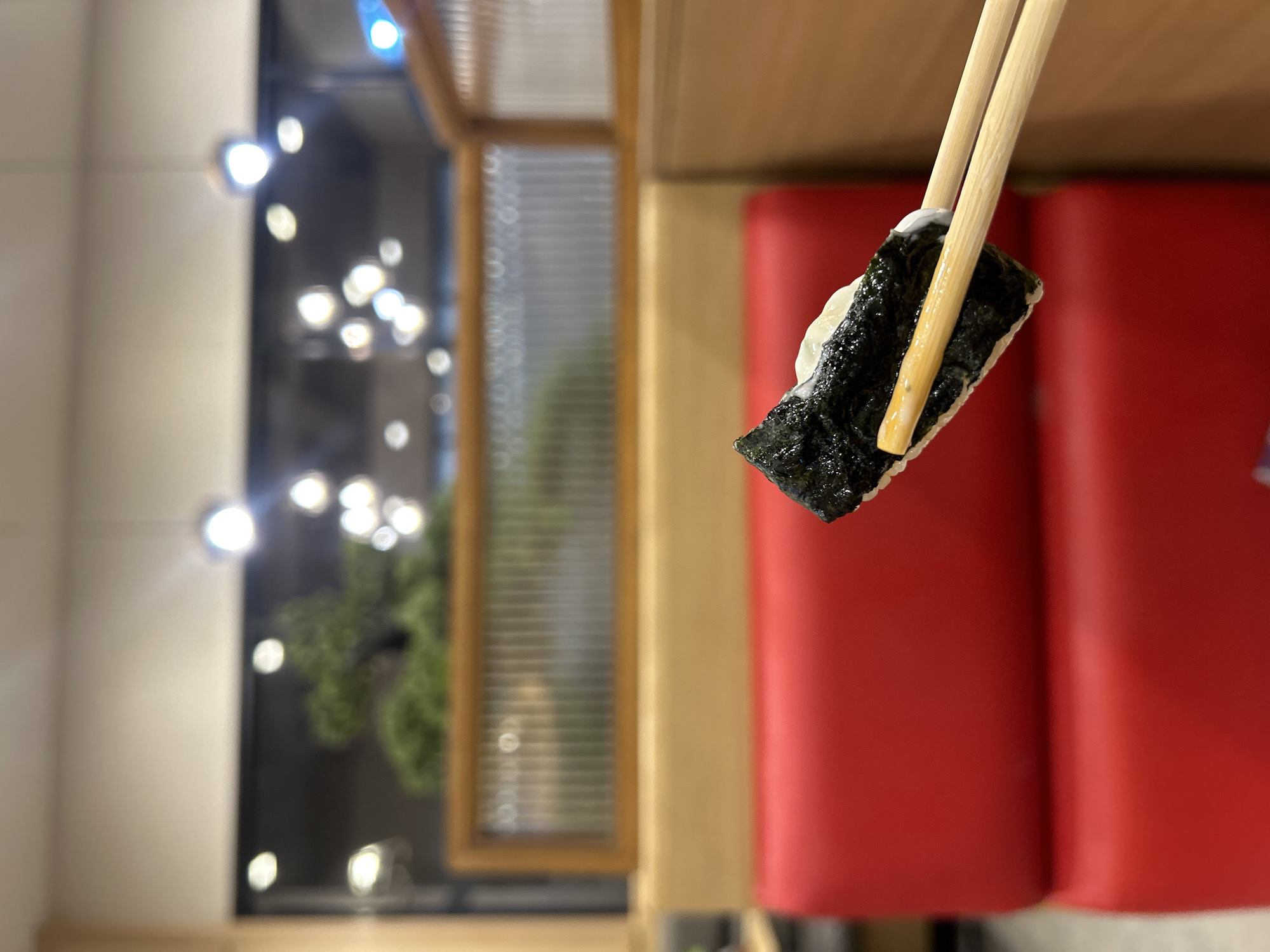 The image shows a close-up of a piece of sushi being held by chopsticks. The sushi is wrapped in nori seaweed and appears to have rice and some white filling. The background is blurred, showing a dining area with red booths and wooden accents, along with some decorative lighting. The overall scene suggests a restaurant setting. There is no information in the image that allows for the identification of a specific location within Tha Khlong Town Municipality, Thailand.