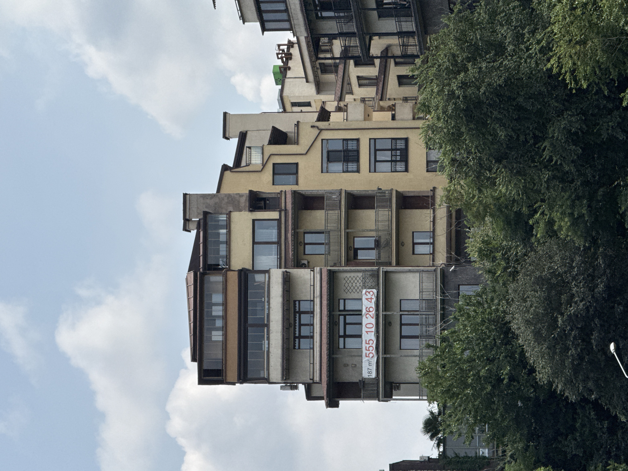 The image shows a multi-story apartment building with a prominent "For Sale" banner featuring a phone number. The banner reads "187 m² 555 10 26 43". The building's architecture is varied, with different sections having distinct styles and colors. Some parts are cream-colored, while others are yellow or have glass enclosures. There are multiple balconies and windows visible. The building is surrounded by lush green trees and foliage in the foreground and midground, obscuring the lower levels and the ground. The sky above is partly cloudy. The scene appears to be a residential area in Tbilisi, Georgia, based on the architectural style and the presence of a real estate advertisement.