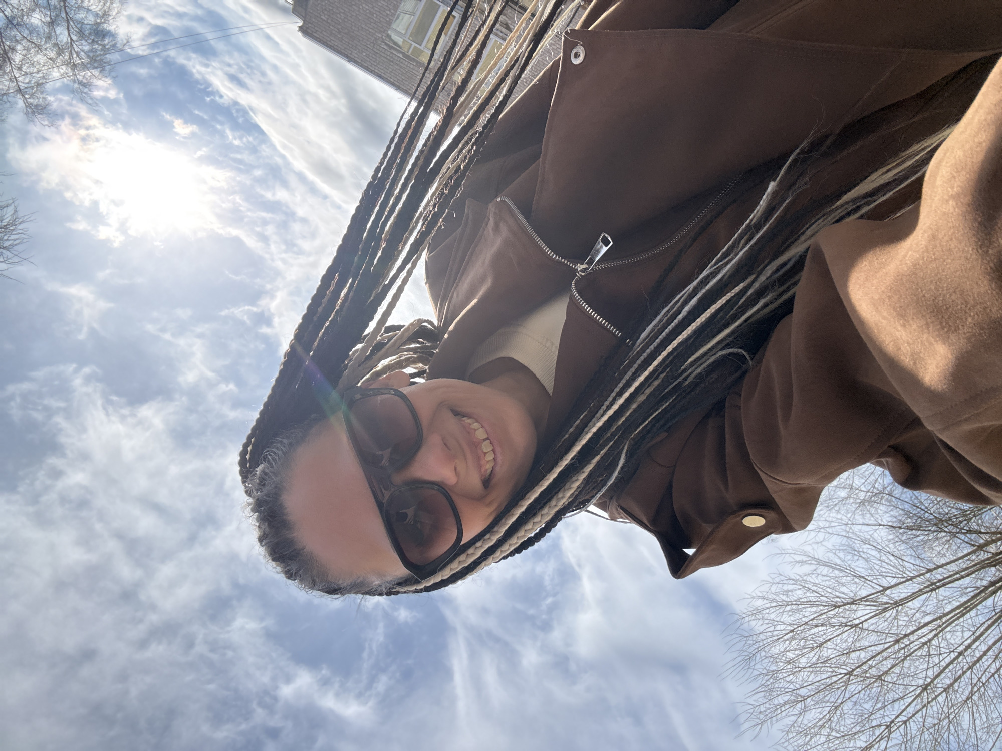 A selfie-style photograph, taken from a low angle, shows a person with long, braided hair with a mix of dark and light strands, wearing sunglasses, and a brown suede jacket. They are smiling and looking up at the camera. The background is predominantly a cloudy sky with the sun shining through, and branches of trees are visible in the bottom right corner. A building can be seen in the top right corner of the image. The photograph appears to have been taken outdoors in a well-lit environment. There is no immediate indication that the location is Rusanovo, Russia, without additional information.