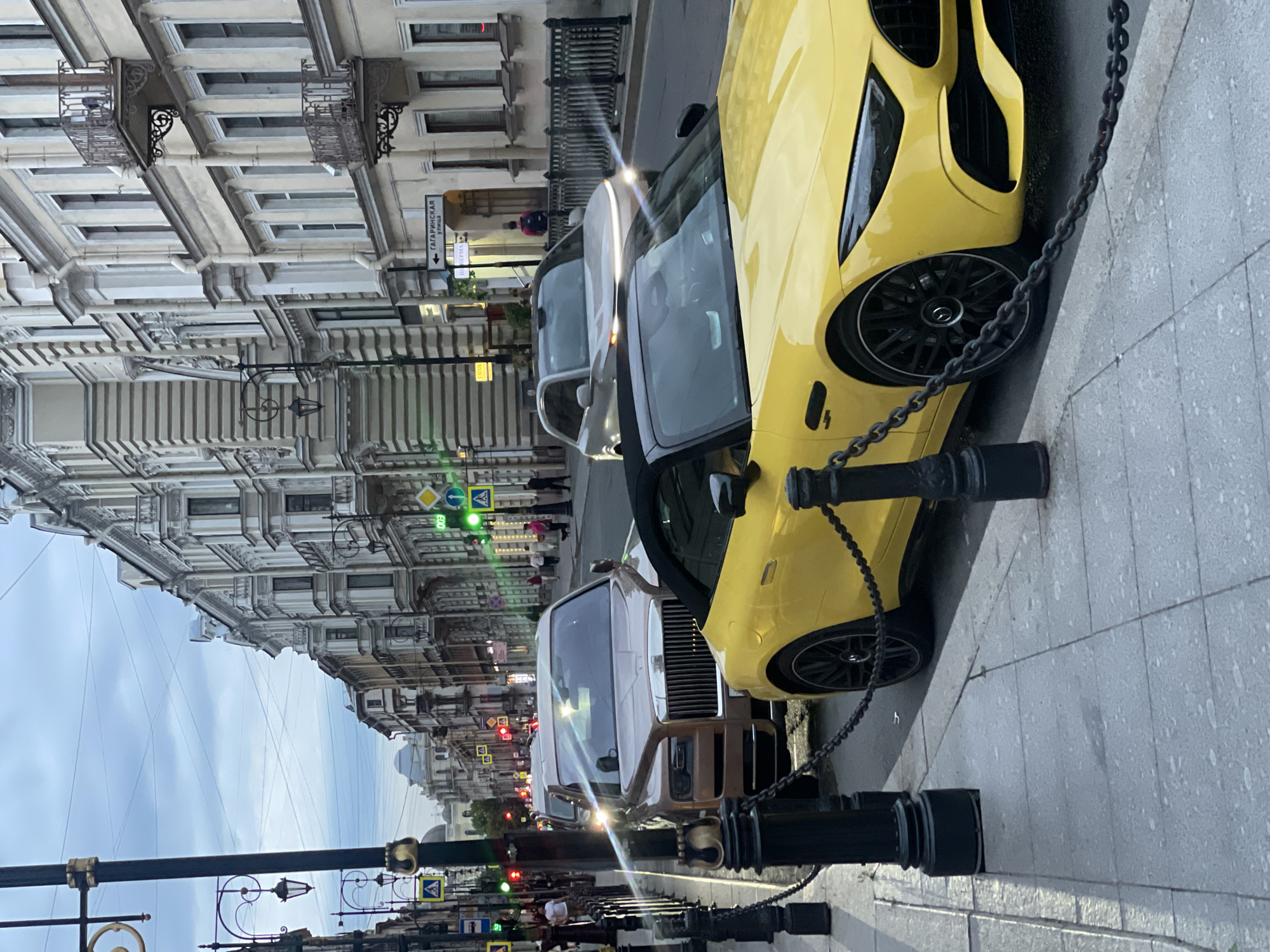 The image shows a street scene in Saint Petersburg, Russia, with several luxury cars parked along the sidewalk. In the foreground, a bright yellow convertible sports car is parked next to a large, tan Rolls-Royce. Both cars are parked on a cobblestone street, and a black metal chain links bollards that line the sidewalk. In the background, older European-style buildings with ornate architecture line the street. Traffic lights and street lamps are visible, and pedestrians are walking along the sidewalk. The sky is overcast.