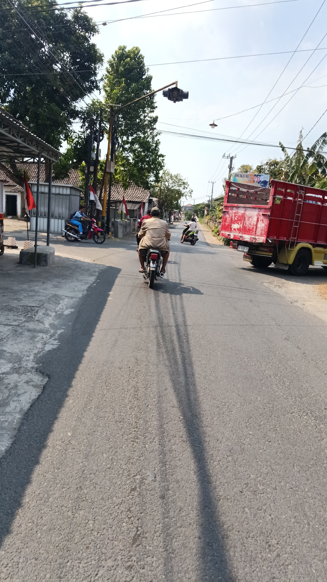 In Dukuh, Indonesia, on a sunny day, a person rides a motorcycle down a paved road. The rider is wearing a light-colored, loose-fitting shirt and dark shorts. In front of them, another person on a motorcycle is also riding. To the right of the road, a large red truck with a yellow cab is parked. The surrounding environment includes trees, buildings with red and white flags displayed, power lines, and traffic signals. The road is casting long shadows, indicating the time of day.