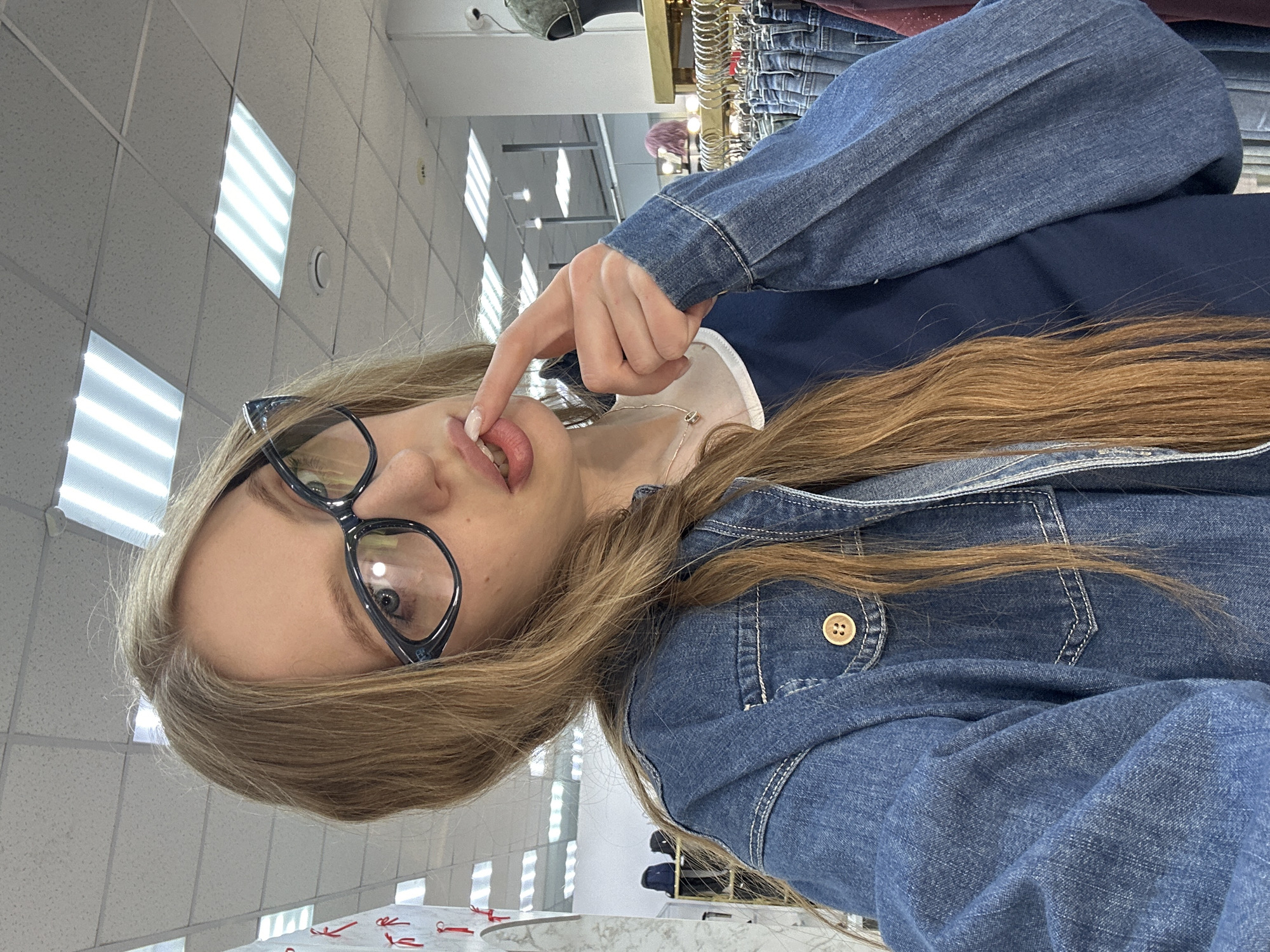 A fair-skinned, blonde-haired woman wearing eyeglasses, a navy blue shirt with a white collar, and a denim jacket is holding her finger to her lips. She is positioned in a retail environment, possibly a clothing store, in Chaikovsky, Russia, as evidenced by the overhead fluorescent lighting and the clothing racks visible in the background. The countertop below her is marble-patterned with red tags.