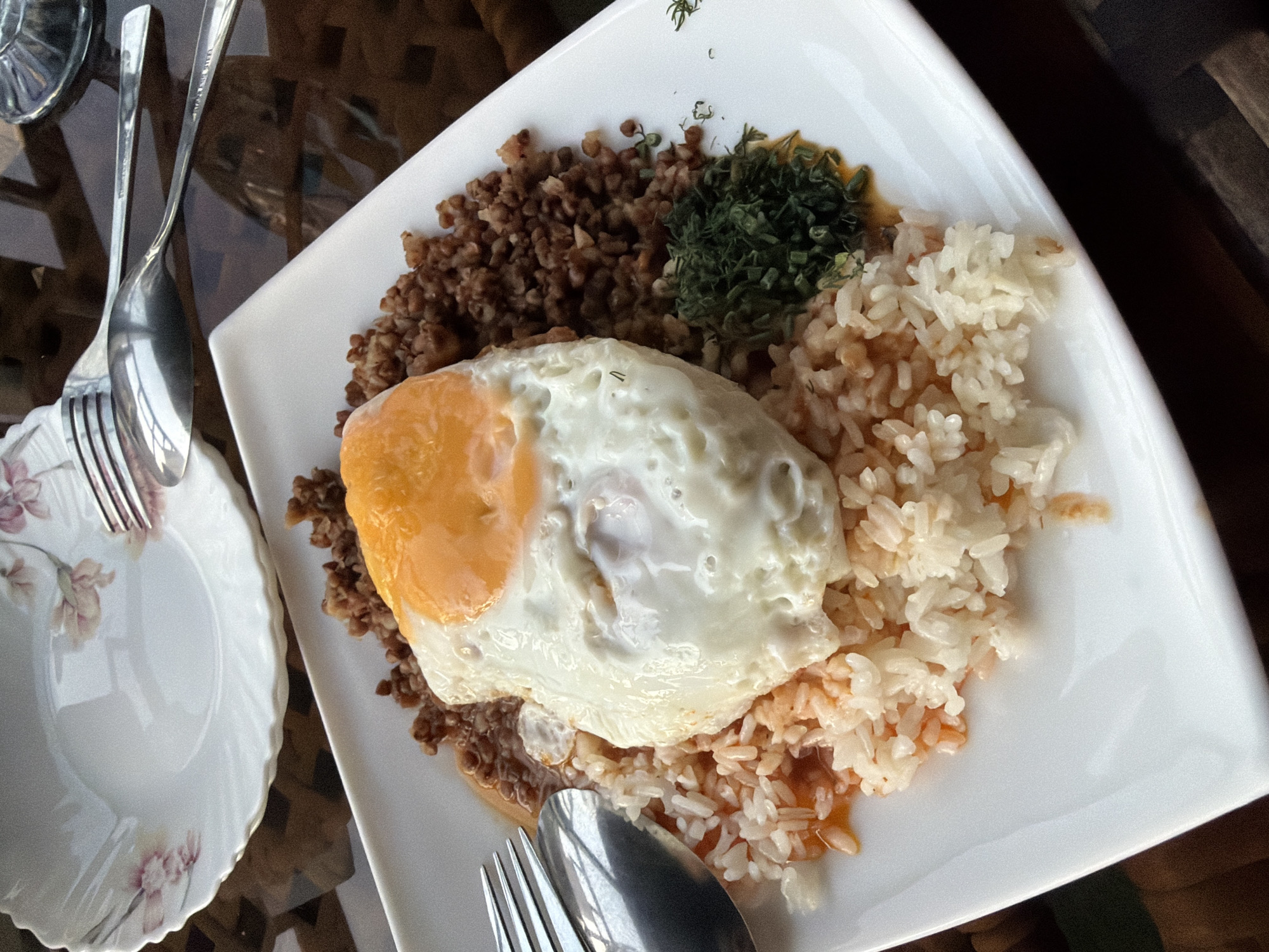 An overhead, close-up shot shows a white plate with a meal on a dark, reflective surface. The meal consists of a fried egg with a bright orange yolk and white albumen placed on top of a bed of rice. To the left of the rice, a portion of buckwheat is visible, with a small pile of chopped green herbs on the right. A fork and spoon are positioned on the upper left of the plate. In the background, another white plate with a floral pattern is visible. The scene is indoors, and there is no indication of the city of Nanay, Uzbekistan.