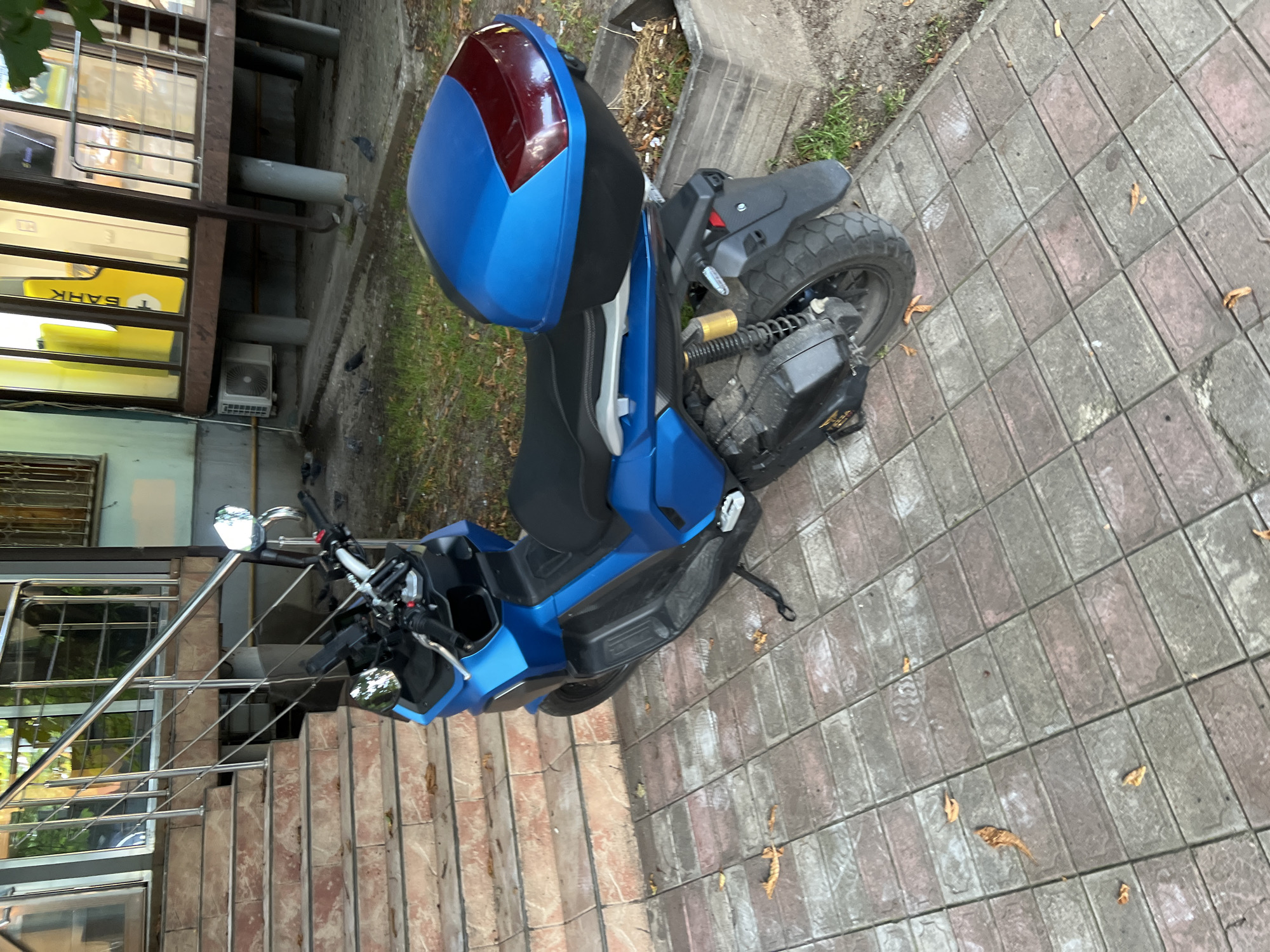 A blue scooter with a red and blue top box is parked on a tiled walkway next to a building. The building has large windows with metal grates and a sign with yellow text that reads "БАНК" (Bank). There are also stairs with a metal railing leading up to the building. The ground is covered in rectangular tiles, and there are fallen leaves scattered around. The time of day appears to be daytime, possibly overcast, given the lighting. The scene is set in Krasnodar, Russia. There are no people visible in the image.
