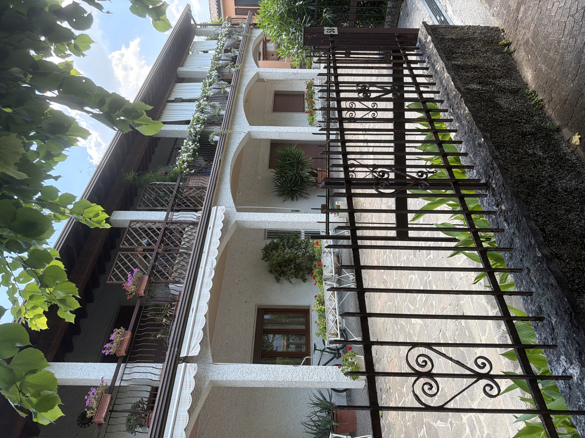 The image shows the exterior of a multi-story building with balconies, located in Padenghe sul Garda, Italy. The building has a white stucco exterior and wooden accents. The balconies are adorned with numerous potted plants and flowers, with a dark metal railing. White curtains hang from the upper balcony, and awnings are visible on the lower level. In front of the building, a dark metal fence encloses a paved courtyard area. A bicycle is parked near a window on the ground floor. Lush green foliage from trees overhangs the top of the image, with a partly cloudy blue sky visible in the background. The overall scene depicts a residential property in a well-maintained state.