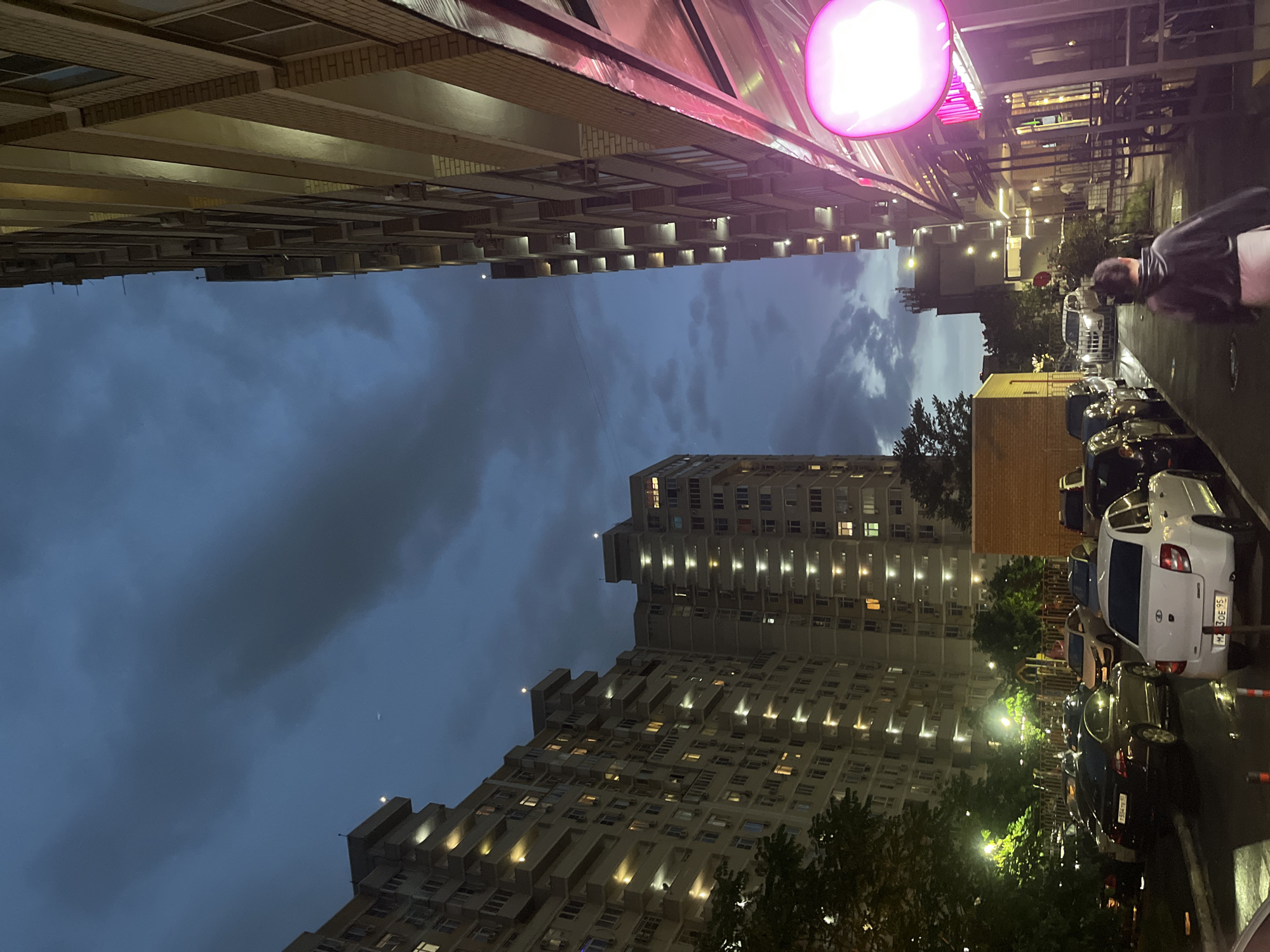 This is a nighttime urban scene in Krasnodar, Russia. The photo is taken from street level, looking upwards at tall apartment buildings on either side, creating a narrow view of the sky. The sky is dark blue with some clouds. The buildings are illuminated with interior lights from the apartments, and some exterior lighting. Parked cars line the street on the right side. A person in a dark jacket is partially visible in the upper right. The focus is on the architectural details of the buildings and the urban environment. A pink, illuminated sign is visible above.