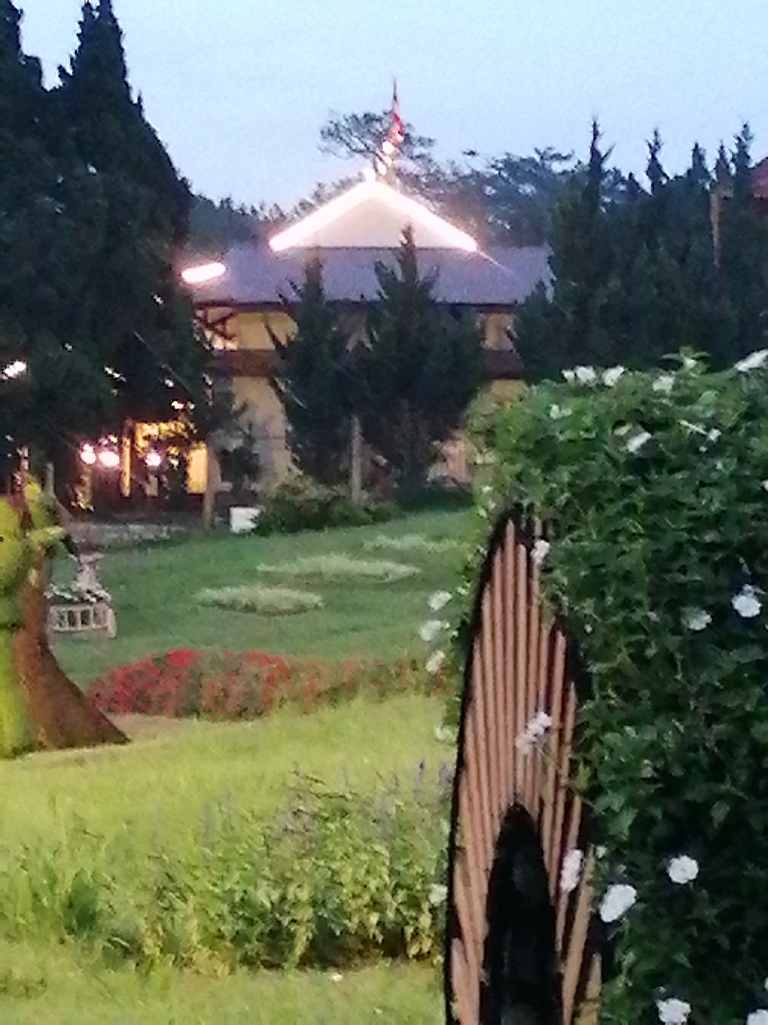 The image shows a garden area with a building in the background, possibly part of a resort or hotel, located in the city of Da Lat, Vietnam. The building has a dark roof with illuminated decorative lights. In the foreground, there is lush green grass, flowerbeds with red flowers, and several trees. A decorative wooden structure with vertical slats is visible on the right side, partially obscuring a dense green bush with white flowers. In the mid-ground, there is a well-maintained lawn with flower arrangements and a decorative element that appears to be a small cart. The overall scene suggests a well-landscaped outdoor space, likely a tourist attraction or a private residence in Da Lat, a city known for its pleasant climate and gardens. The sky is overcast, indicating it might be late afternoon or early evening.