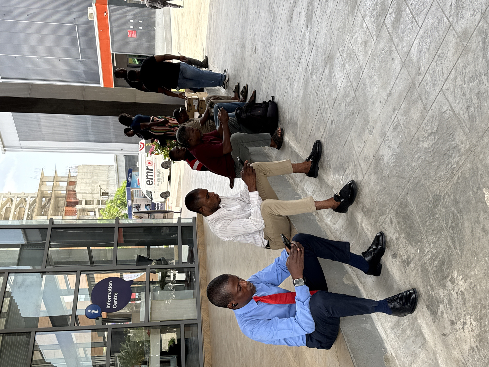 A group of men sit on the pavement outside an "Information Centre" in Alaguntan, Nigeria. They are oriented in a line, with varying degrees of formality in their attire, most appearing to be using their phones. A building under construction is visible in the background, as well as a vehicle marked "emro". The pavement is concrete with a patterned surface.
