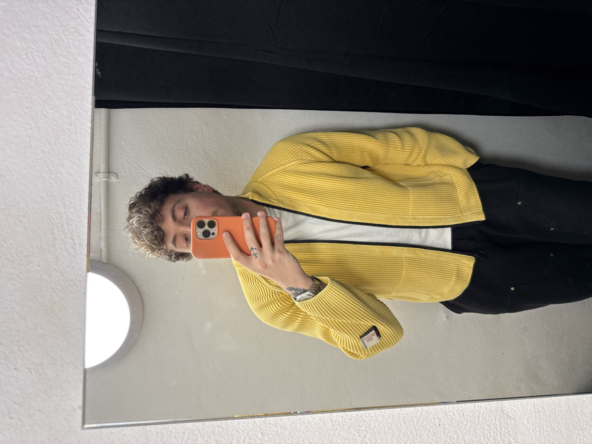 A young man is depicted taking a mirror selfie indoors, holding an orange smartphone. The image is oriented horizontally, showing the man's reflection as if he is standing upright. He has curly dark hair with lighter tips and is looking down at his phone screen.
He is wearing a bright yellow, ribbed zip-up jacket over a white t-shirt and black trousers. On the lower left sleeve of his jacket, there is a small rectangular patch with text that appears to be "NOKOO" or "NOKOD" and a small logo. He has a silver watch on his left wrist and tattoos on his left hand (an anchor) and forearm.
The background in the mirror reflection consists of a plain white wall and a dark black fabric or curtain. A circular light fixture is partially visible in the top left of the mirror's reflection. The wall where the mirror is mounted has a textured white surface. The setting suggests an indoor space, possibly a fitting room, located in London, United Kingdom.