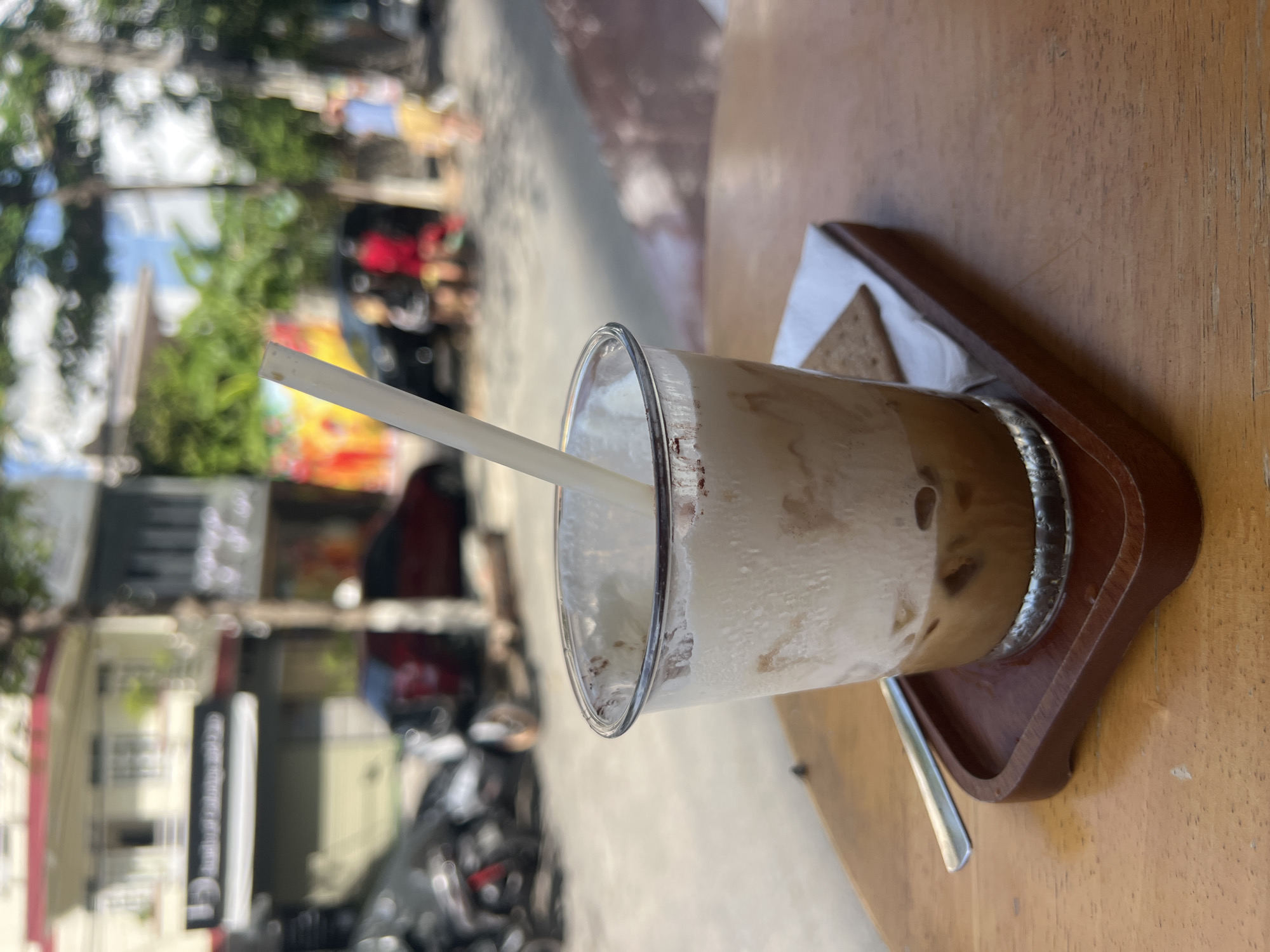A close-up shot shows a clear glass of iced coffee with a thick layer of whipped cream on top, a white straw inserted into it, and a small square cracker on a white napkin, all resting on a wooden tray. The tray is placed on a wooden table. In the background, a street scene unfolds with blurred buildings, trees, parked motorcycles, and indistinct figures of people walking or standing. The lighting suggests it is daytime, possibly afternoon, with sunlight casting shadows.