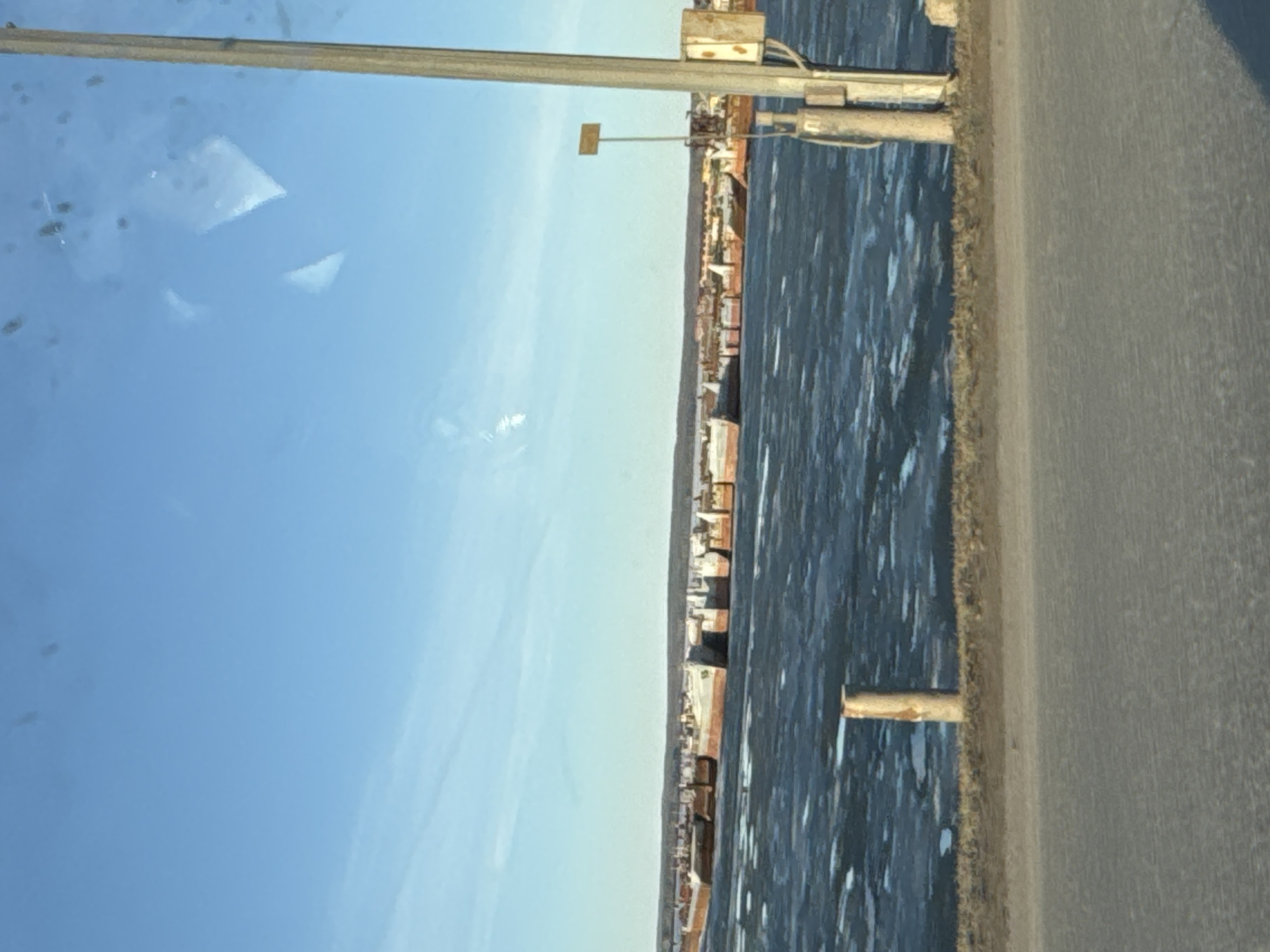 The image shows a view from a vehicle (possibly a car or bus) in Chaikovsky, Russia. The sky is a clear, light blue with a few faint clouds visible. A metal pole with some equipment attached runs along the upper portion of the frame. To the right, a body of water, possibly the Kama River, stretches out toward a shoreline dotted with buildings, indicating the edge of the city. A road or highway is visible on the right side of the image, with a gray surface. The image is slightly tilted.