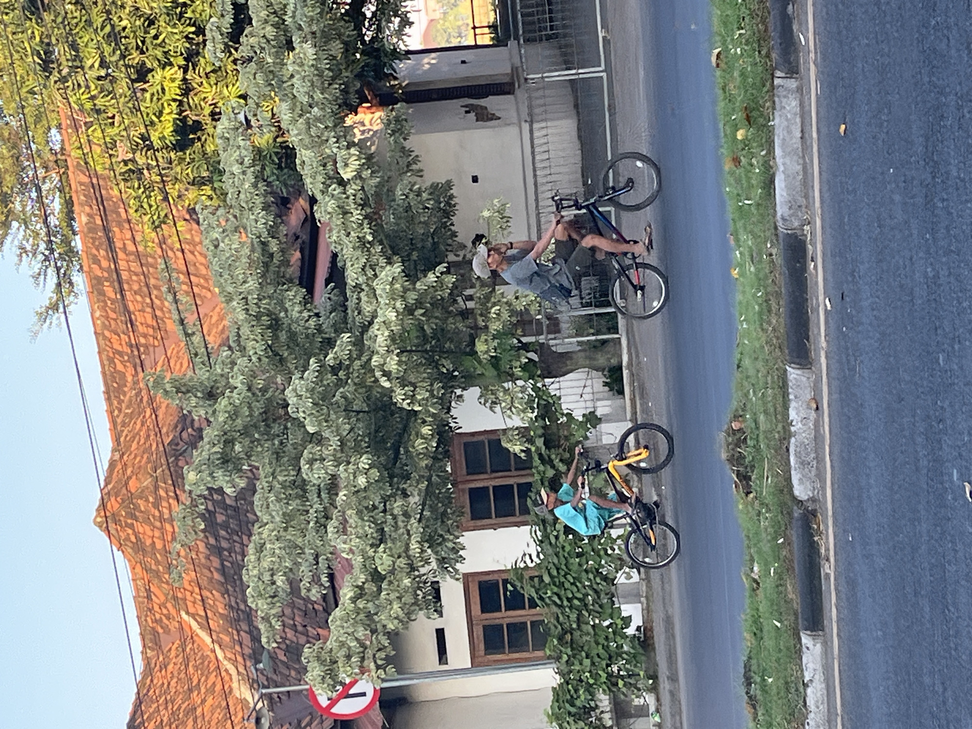 Two individuals are cycling on a street in Kludan, Indonesia. The person on the left is a young boy wearing a blue shirt and shorts, riding a yellow and black bicycle. The person on the right is an adult male wearing a gray t-shirt, shorts, and a cap, riding a multicolored bicycle. They are both in motion, on a paved road with a grassy area and a curb on the left side. Behind them is a building with a tiled roof and large windows, surrounded by trees and foliage. A "no entry" sign is visible on the left. The overall scene depicts a daily activity in a residential or commercial area of Kludan.