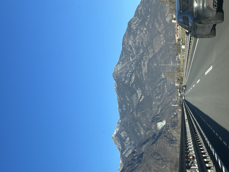 This image captures a bright, clear day along a multi-lane highway in a mountainous region, consistent with the landscape of Verrès, Italy. The prominent subject is a vast, rugged mountain range dominating the left side of the frame, showcasing dark rock faces and scattered lighter patches, possibly snow or lighter rock formations. The highway occupies the right side, extending into the distance, with distinct white lane markings and a metal guardrail running parallel to the mountain.
In the upper right foreground, a dark-colored car, possibly an SUV, is partially visible, indicating the photo was taken from a moving vehicle. Other vehicles appear as small dots in the distance on the highway. Along the base of the mountain, some indistinct buildings and infrastructure can be seen, suggesting a mix of natural landscape and human presence. The sky is a cloudless, vibrant blue, indicating fair weather and bright daylight. There is no discernible text visible in the image. The scene suggests travel through a scenic alpine environment.