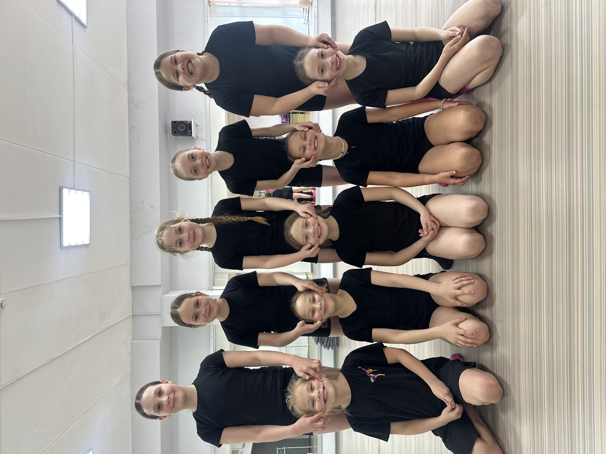This image captures nine young girls, likely ranging from pre-teen to early teen age, engaged in a playful activity indoors. They are arranged in a vertical line, forming a human pyramid-like structure, with each girl sitting or kneeling behind and slightly above the one below her. All subjects are dressed uniformly in black short-sleeved t-shirts and black shorts, and most have their hair styled in braids or ponytails.
The central activity shows a chain of interaction: each girl, starting from the second one from the top, is using both hands to gently hold and stretch the cheeks of the girl in front of her, creating exaggerated, wide smiles. All the girls in the photograph are actively participating, displaying joyful and amused expressions, characterized by wide smiles and twinkling eyes, suggesting a moment of fun and camaraderie.
The setting appears to be a brightly lit indoor space, possibly a dance studio, gym, or activity room. The floor is light-colored, featuring subtle horizontal stripes, while the walls are plain light grey or white. Rectangular LED light fixtures are visible on the ceiling/wall. A single black speaker is mounted on the wall in the upper right of the frame. The atmosphere conveyed is cheerful and lighthearted. The image captures a daytime scene, given the interior lighting. No discernible text is visible on clothing or walls. This scene took place in Votkinsk, Russia.