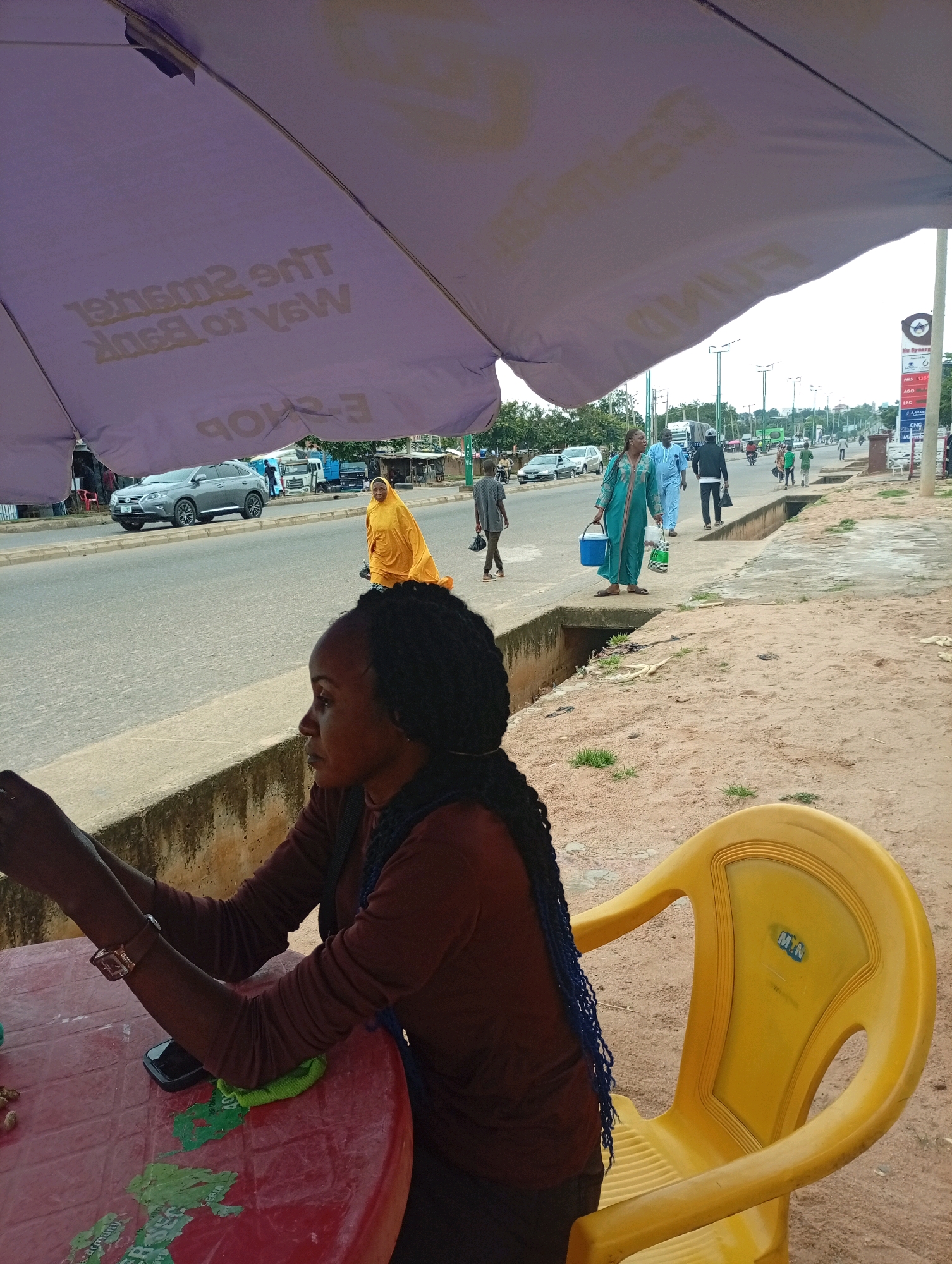 An eye-level, medium shot captures a woman seated at a red, round table under a large, purple umbrella. The woman, with dark skin and braided hair, wears a brown long-sleeved shirt and is looking to her left. A dark-colored phone lies on the table next to a green cloth. To her right, a bright yellow plastic chair with the "Mtn" logo is visible. The background shows a street scene in Garko, Nigeria, with a road, cars, and pedestrians. A gray SUV is parked on the left, while several people walk along the sidewalk on the right. A tall, green pole with streetlights is present on the sidewalk, and a sign with multiple fuel prices is visible in the distance. The ground is covered in sand and dirt.