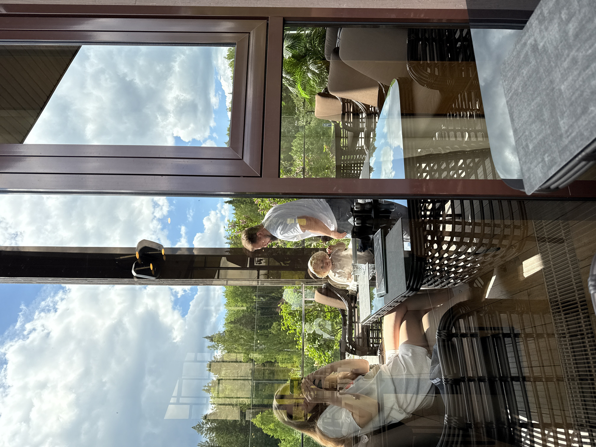 The image presents a bright daytime scene viewed through a large window, characterized by complex reflections that superimpose interior and exterior elements. The dominant background is a clear blue sky with fluffy white clouds, seen through and reflected on the windowpanes. Below the sky, a lush landscape of green trees and foliage is visible, suggesting a natural outdoor setting in Dobrograd, Russia.
Inside, the setting appears to be a modern and comfortable space, possibly a cafe or restaurant. Three individuals are present: a man in a light-colored t-shirt stands near a table, looking towards a small child with light hair who is seated. Further right, a woman is seated, with her hands near her face, possibly holding a phone. They are surrounded by contemporary, wicker-style furniture including chairs and a table. Reflections on the glass create a layered effect, showing the indoor scene and furniture overlaid with the outdoor environment. An electronic device, possibly a speaker or camera, is mounted on the interior window frame. The overall atmosphere is relaxed, with bright natural light suggesting midday or early afternoon. No clear text is visible.