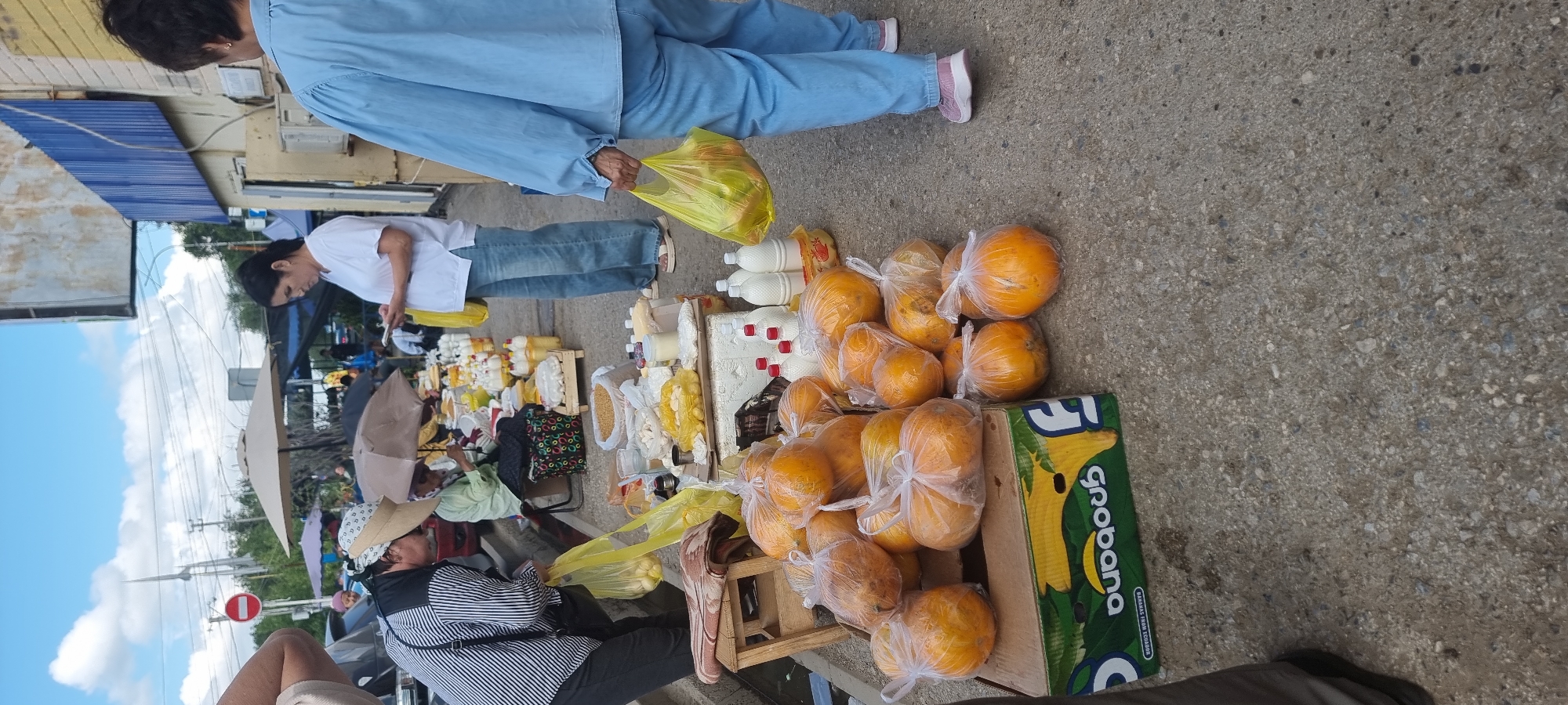 In Aktobe, Kazakhstan, an outdoor market scene unfolds. A woman in a white t-shirt and blue jeans stands looking at her phone, while another woman in a blue jumpsuit carries a yellow plastic bag filled with produce. In the foreground, an assortment of round, orange fruits, likely melons or pumpkins, are bagged in clear plastic and arranged on a green and white cardboard box. Other food items, including what appear to be dairy products in bottles and containers of grain, are displayed on tables and stands behind the fruit. The market is set up along a paved walkway, with various umbrellas providing shade for the vendors. The backdrop includes a building with a light-colored facade and blue trim, suggesting an urban setting.