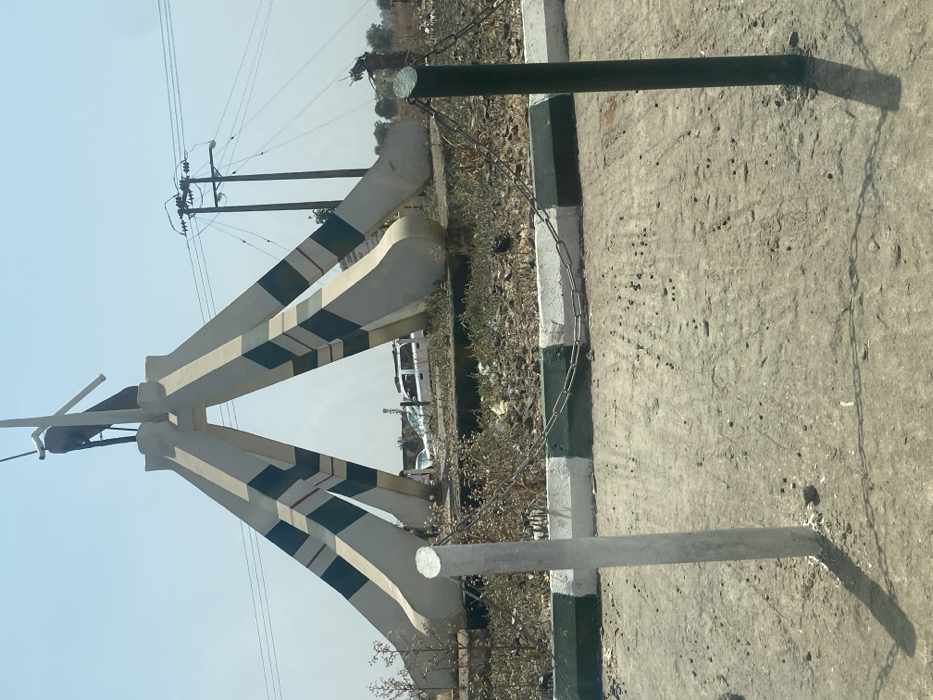 This image displays a street scene in Unguwar Gaya, Nigeria, under bright, clear daylight conditions. The central subject is a monument featuring four robust, upward-tapering concrete pillars painted white with horizontal green and thin red stripes, converging at an apex. A dark, abstract figure, possibly avian or mechanical, is mounted at the monument's peak.
In the immediate foreground, a light-colored, uneven dirt surface extends across the lower frame. A white vertical post is situated on the left, and a dark green/black vertical post is on the right. These posts are linked by a single strand of silver-colored metal chain, forming a low barrier. Behind this barrier, a concrete curb, painted with alternating white and dark green sections, defines the edge of a circular central island. This island's surface consists of dry, sparse vegetation, exposed dirt, and scattered debris.
In the background, behind the monument, a white SUV or van is partially visible, alongside another darker vehicle, suggesting a road or parking area. A solitary, indistinct figure is faintly discernible near the white SUV. To the right, multiple wooden utility poles with an array of overhead power lines are visible against a pale blue, cloudless sky. Distant, dry vegetation and minimal structures line the horizon.