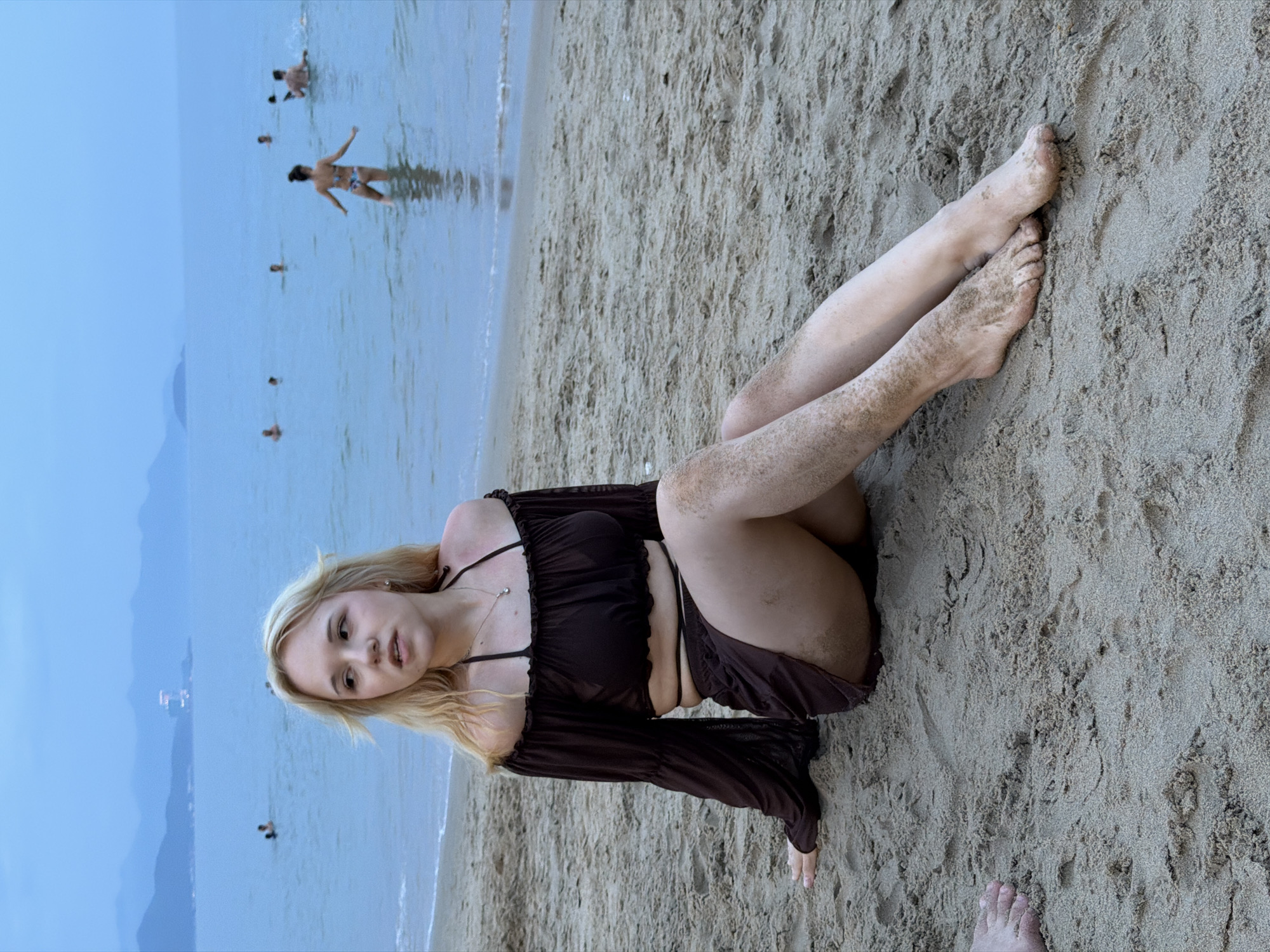 A young Caucasian woman with blonde hair sits on a sandy beach, looking to her left. She is wearing a dark brown, off-the-shoulder crop top with long bell sleeves and a matching skirt. Her legs are partially covered in sand. In the background, the ocean is calm with several people swimming and wading in the water. Distant mountains are visible on the horizon. The sky is overcast with a twilight hue, suggesting it is either early morning or late evening. The location appears to be a beach in Nha Trang, Vietnam, based on the presence of similar landscapes in other images from the city.