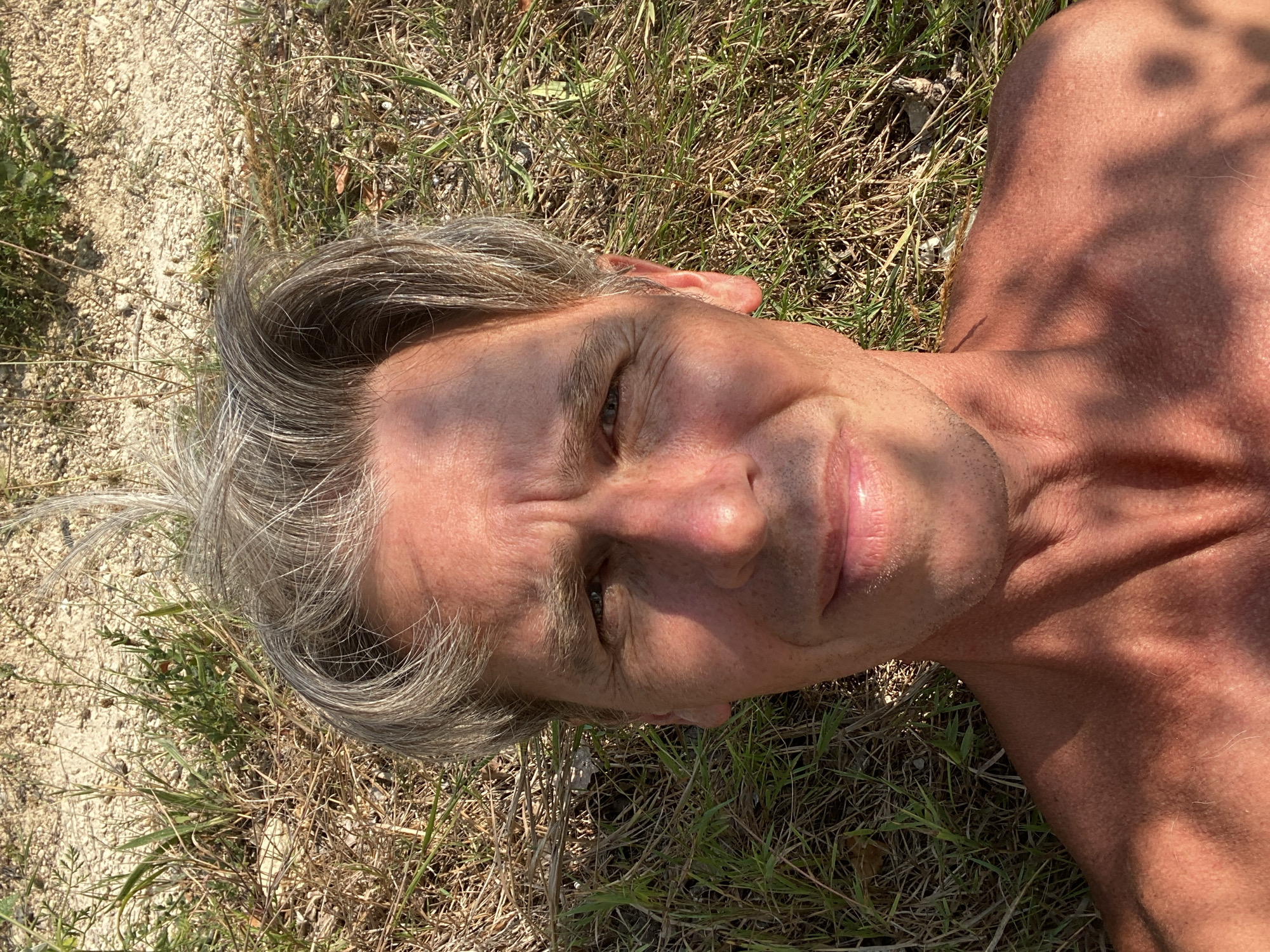 The image is a close-up, upward-facing selfie of a shirtless man lying on the ground outdoors. He has short, graying hair and is looking directly at the camera with a slight smile. His skin is tanned and shows some sun damage. The background consists of dry grass, dirt, and some small stones, suggesting a rural or natural setting, possibly in the city of Kuibyshevske, Ukraine. The lighting indicates it is daytime, with dappled shadows falling across the man's chest and neck.