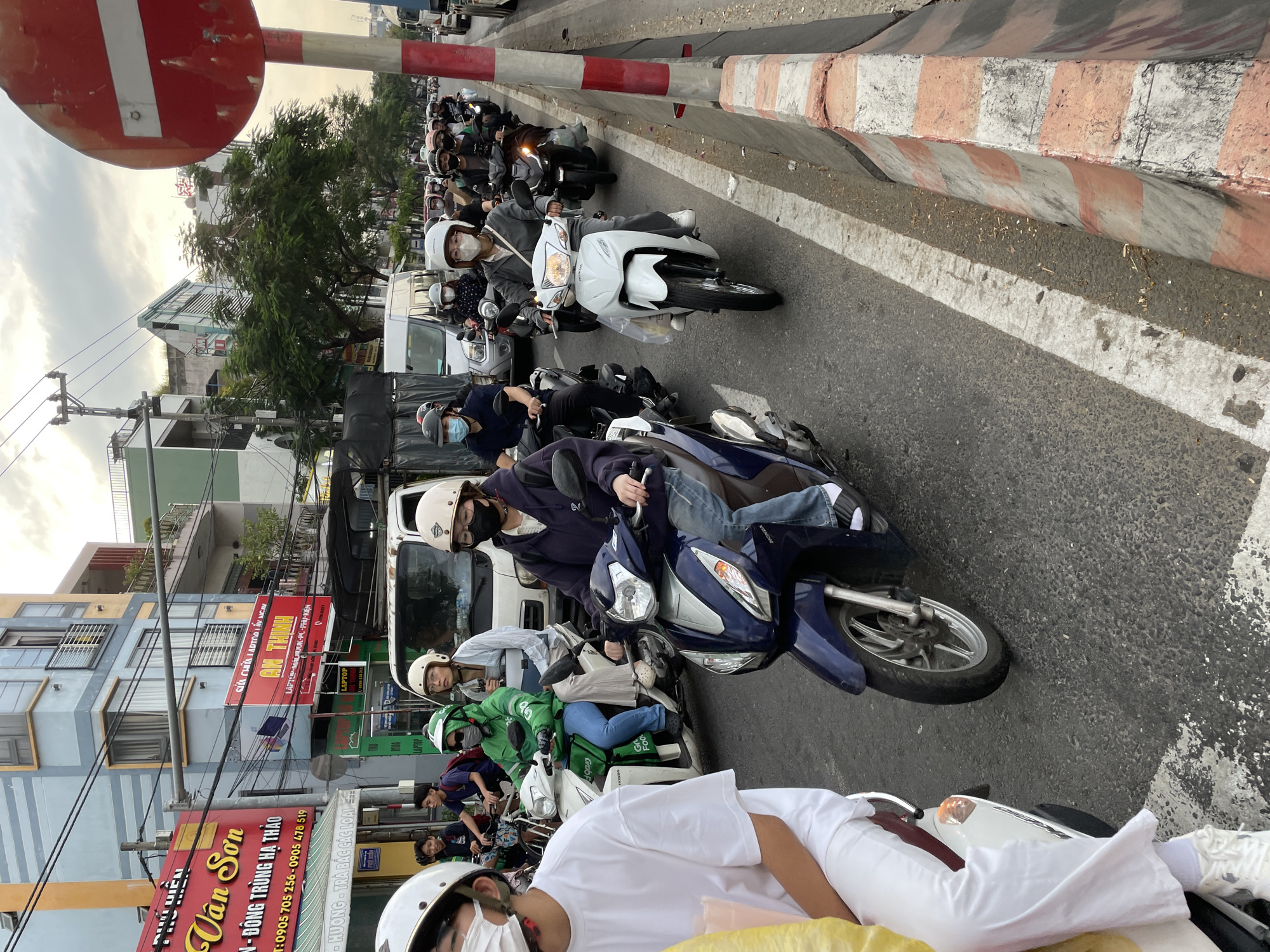 This is a medium shot of a traffic jam in what appears to be a city in Vietnam, possibly Da Nang. The image is taken from the perspective of someone on a motorcycle, looking forward. The primary subjects are numerous people riding motorcycles and scooters, many wearing helmets and face masks. The scene is a busy street with buildings lining the side.
The activity taking place is a traffic jam, with many motorcycles queued up. Some individuals are looking forward, while others are looking towards the camera or to the side. One person in the foreground is wearing a white t-shirt and a white helmet. Another person in the middle ground is wearing a green jacket with "GrabFood" branding, suggesting a delivery service.
Notable details include the time of day, which appears to be late afternoon or early evening based on the lighting and the soft, diffused light in the sky. The weather seems to be cloudy or overcast. The buildings visible have signs with Vietnamese text. Some of these signs are for "Vân Sơn" (selling Đông Trùng Hạ Thảo, a type of mushroom) and "An Thịnh" (selling laptops and computer accessories). There is also a "GrabFood" sign visible. The street has a red and white striped barrier along one side.
The overall mood is one of a typical urban commute in a densely populated area, characterized by the common sight of motorcycle traffic. There are no overt displays of emotion, but the scene suggests a common, everyday occurrence.