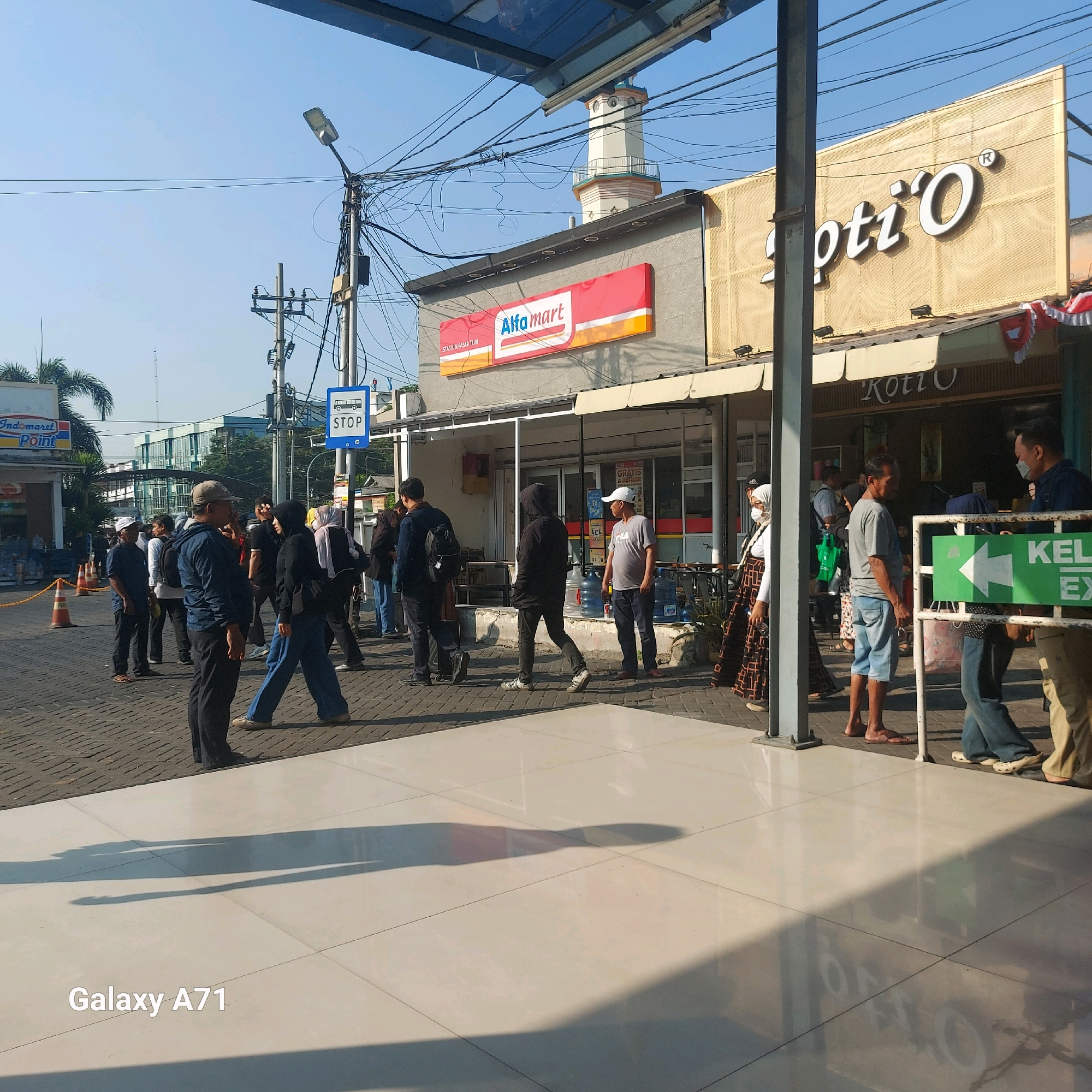 The image captures an outdoor scene in the city of Surabaya, Indonesia, likely on a bright, sunny day with a clear blue sky. The foreground shows a paved walkway with large, square, light-colored tiles, reflecting the sunlight and casting long shadows from an unseen overhead structure. A watermark "Galaxy A71" is present in the bottom left corner.
In the midground, a paved street or sidewalk is visible, lined with people. To the left, a bright orange traffic cone and a yellow barrier suggest a temporary closure or designated area. Adjacent to this is a building with an "Indomaret Point" sign. Further right, a building with a prominent red and white "Alfa mart" sign is visible, with a mosque minaret rising behind it. Next to the Alfa mart, another establishment with a large gold sign featuring white lettering that reads "Koti'O" is present. People are standing in a line or milling about in front of these businesses and a bus stop sign that reads "STOP" with a bus icon. Several individuals are dressed casually, with some wearing masks. There are also a few large water dispensers visible.
In the background, a modern building with glass facades and palm trees are visible, indicating an urban environment. Overhead, a complex network of electrical wires and poles spans the scene, contributing to the bustling streetscape. The overall impression is that of a commercial area with a bus stop, where people are waiting or going about their daily activities.