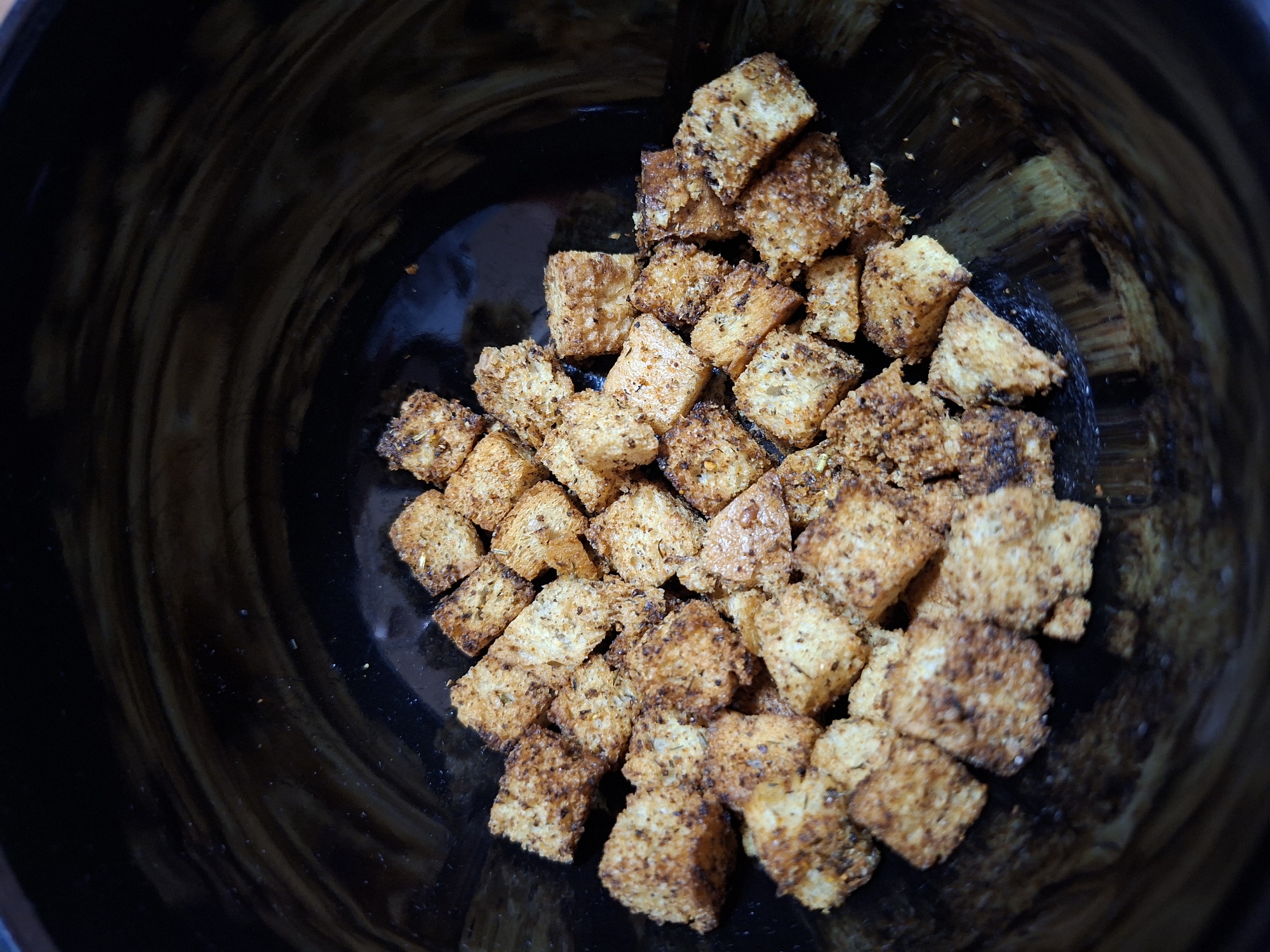 The image displays a close-up, overhead view of a cluster of golden-brown, cube-shaped croutons contained within a dark bowl. The croutons are irregularly piled, showing varying shades of toasted bread and are visibly seasoned with dark specks, suggesting the presence of herbs or spices. The texture appears crispy, with some pieces exhibiting a slightly coarser grain.
The bowl itself is a dark, possibly black or very dark brown, material, possessing a subtle glossy finish. Faint, circular or swirling patterns are visible on the interior surface of the bowl, especially in the areas not covered by the croutons. The background is uniformly dark and out of focus, ensuring the croutons are the central subject.
No specific activity is taking place; the croutons are presented statically, suggesting they are prepared for a meal. There are no people visible, nor are there any direct indications of time of day or weather. While the image itself does not contain location cues, the provided context specifies Plovdiv, Bulgaria. No visible text is present in the image.