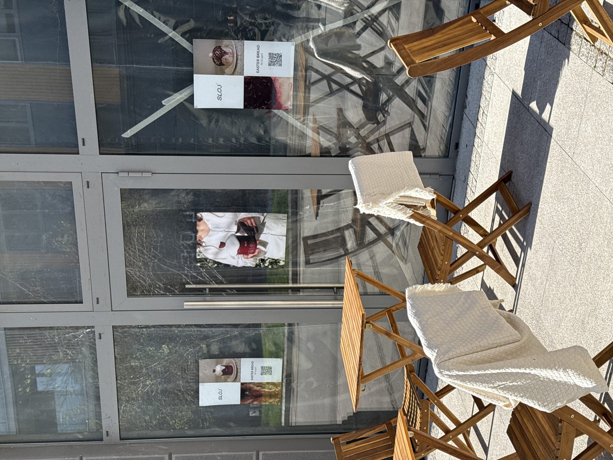 The image shows a cafe in Belgrade, Serbia, from an external perspective. The cafe features a glass-fronted building with posters on the glass advertising "Easter Bread" from "SLOJ". Outside, there are wooden folding chairs with white waffle-textured throws or blankets placed on them, presumably for customer comfort. A small wooden table is also present. The scene is well-lit, suggesting it is daytime. The background reflection in the glass shows trees and some blurred building details, indicating a city environment. No people are directly visible.